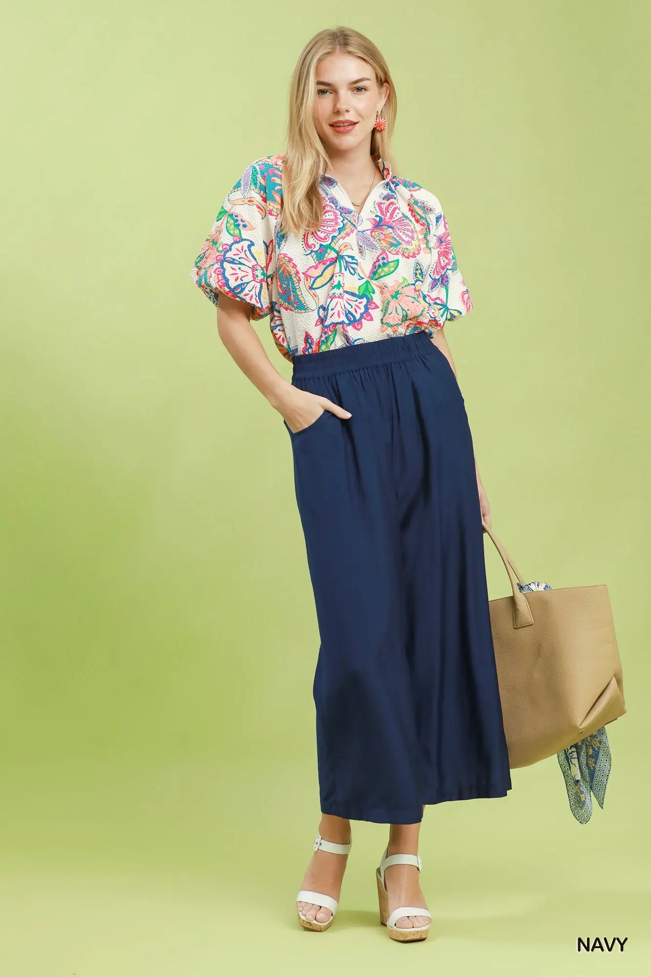 Woman wearing a floral blouse and navy pants with a beige handbag on a light green background