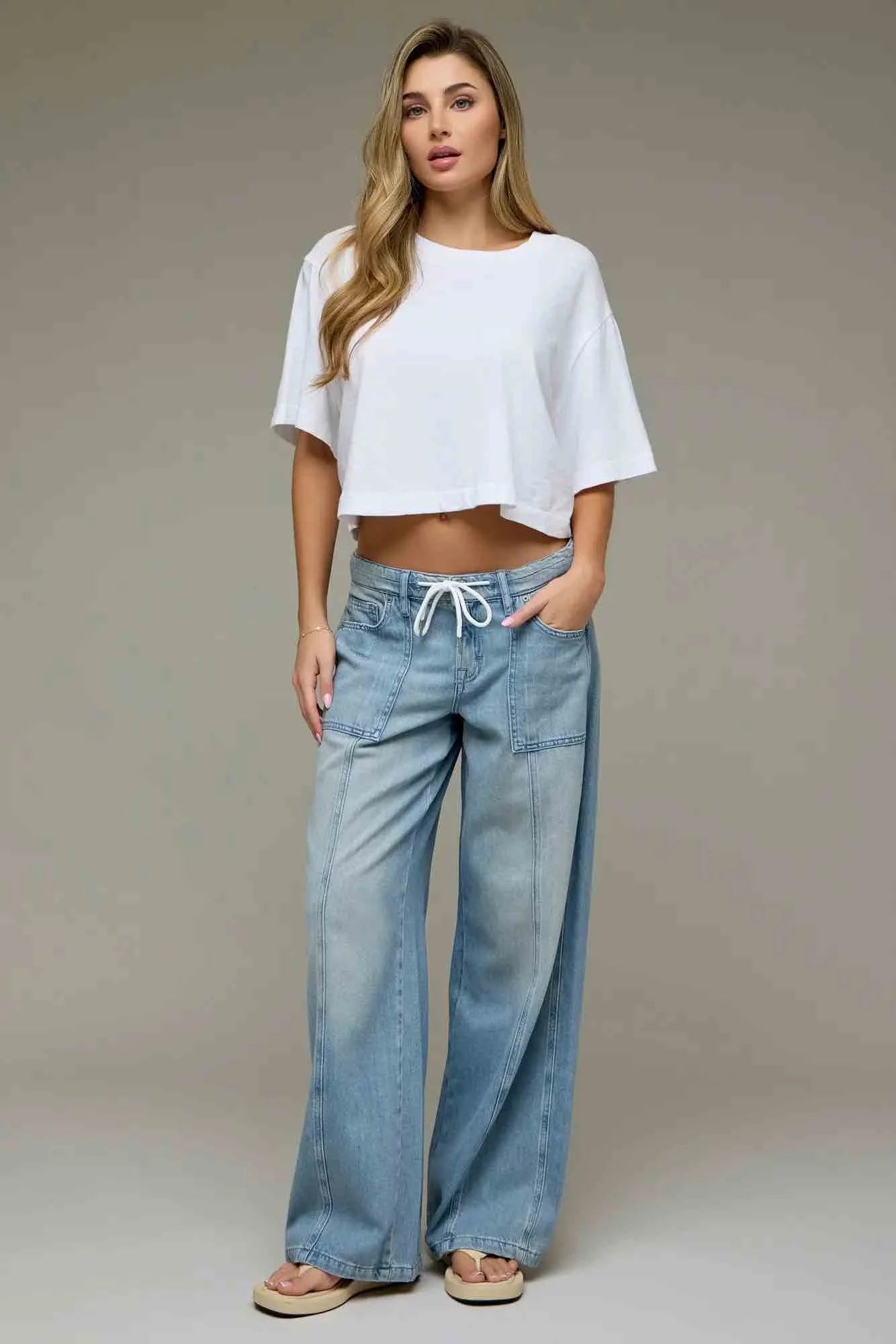 A woman modeling a white cropped short-sleeve t-shirt and light-wash wide-leg denim jeans with a drawstring waist, standing against a neutral studio background.