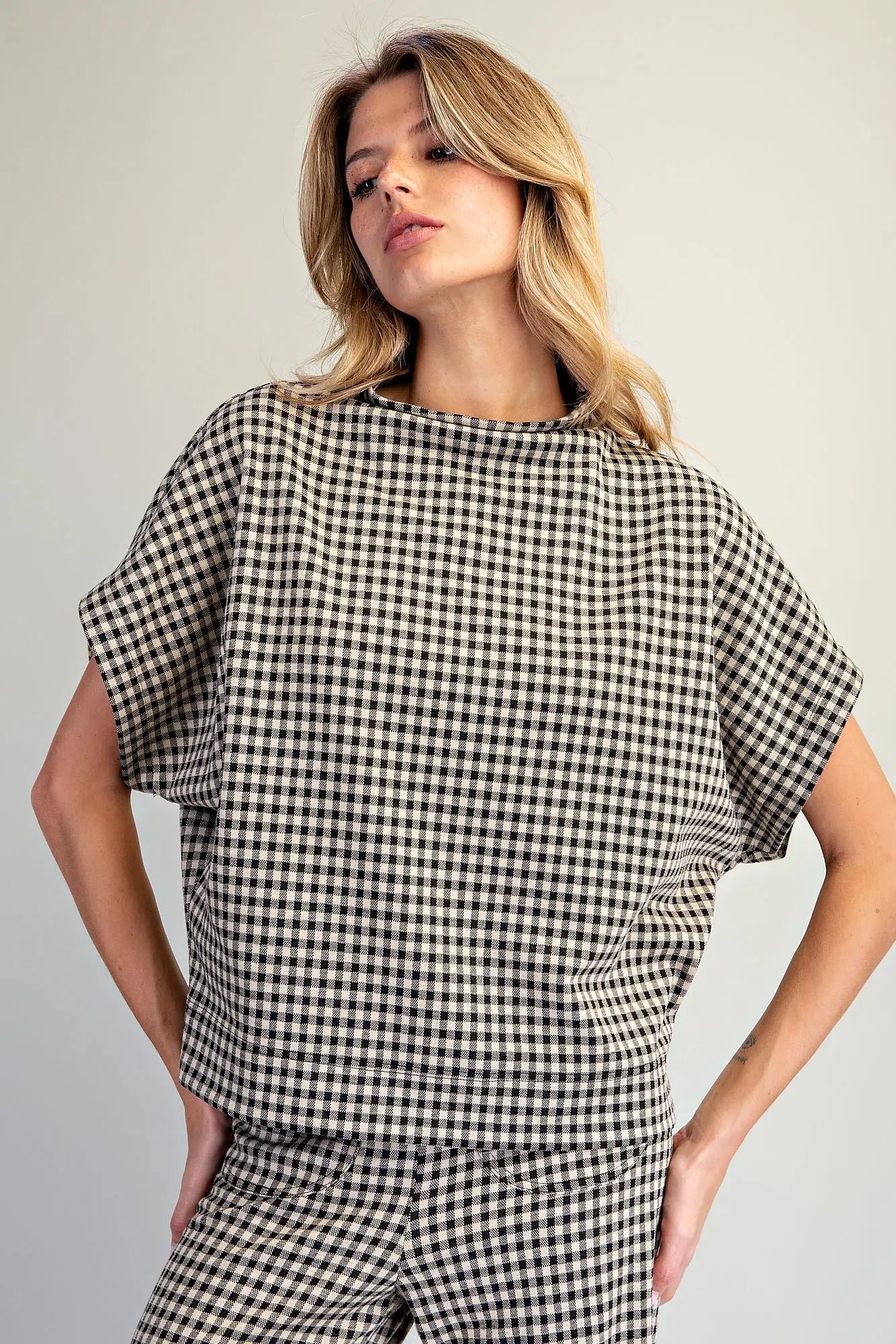 A woman modeling a black and beige gingham check short-sleeve top and matching pants against a plain light gray studio background.