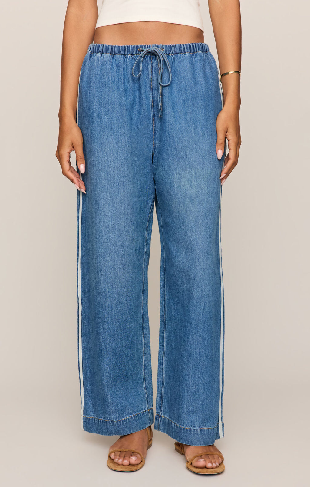 A model wearing a pair of wide-leg blue denim pants with white side stripes, paired with a white crop top and tan flat sandals, against a solid light beige background.