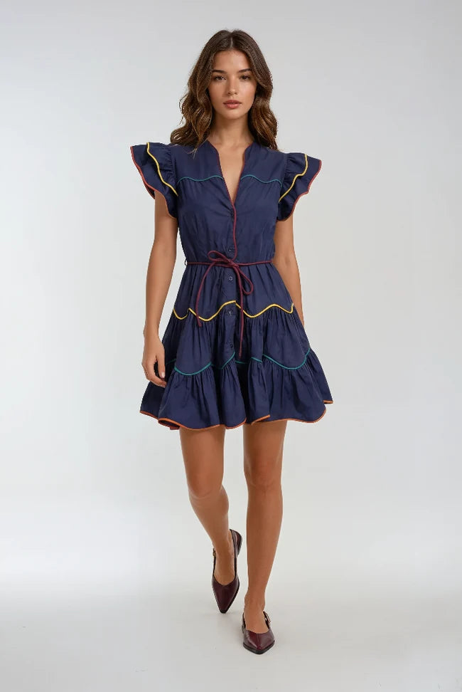 A woman modeling a navy blue mini dress with ruffled cap sleeves, a tie waist, and colorful embroidered trim in yellow, green, and red, set against a plain light gray studio background.