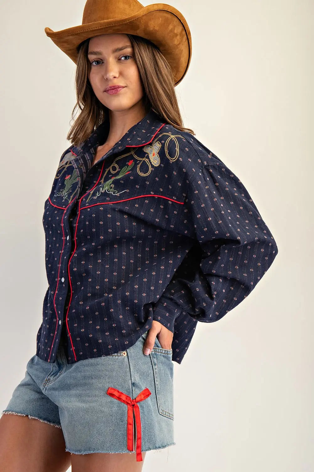 Woman wearing a navy blue embroidered shirt and denim shorts with a brown cowboy hat on a white background