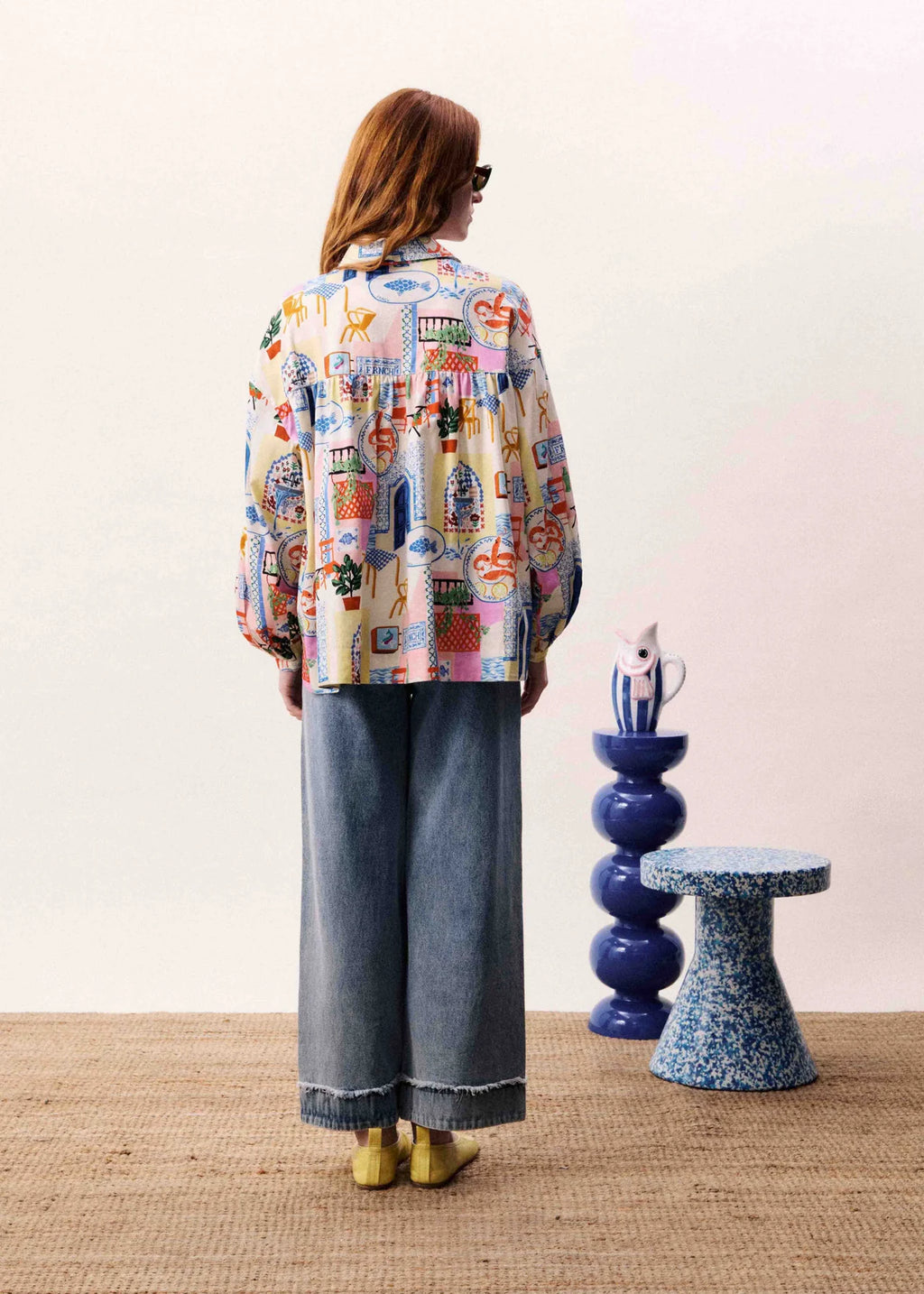 Person wearing a colorful patterned shirt and jeans standing in a room with decorative stools.