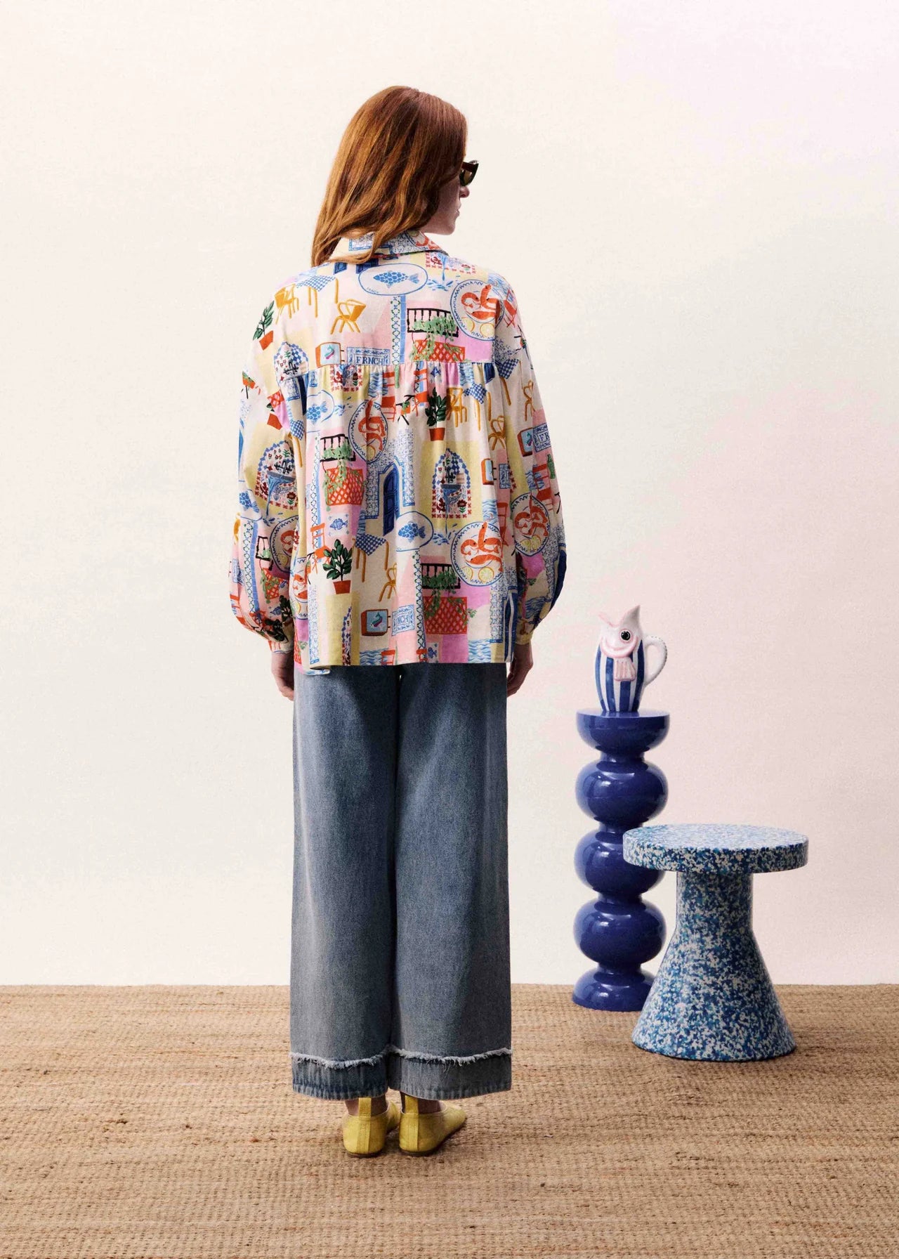 Person wearing a colorful patterned shirt and jeans standing in a room with decorative stools.