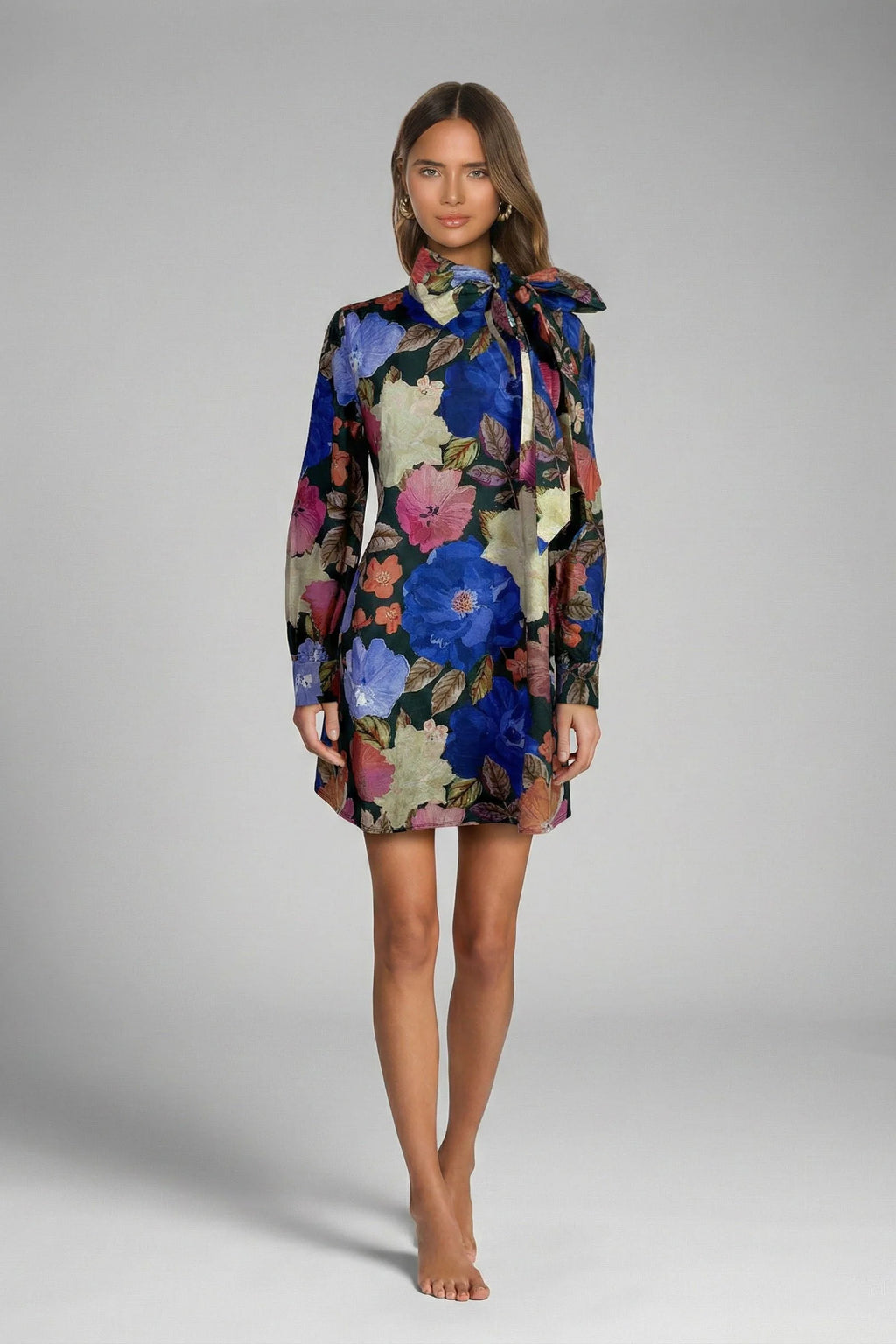 A woman modeling a long-sleeve mini dress with a high neck and bow detail, featuring a vibrant floral print in blue, pink, and beige on a dark background, set against a plain gray studio backdrop.