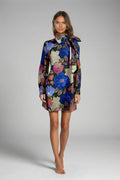 A woman modeling a long-sleeve mini dress with a high neck and bow detail, featuring a vibrant floral print in blue, pink, and beige on a dark background, set against a plain gray studio backdrop.