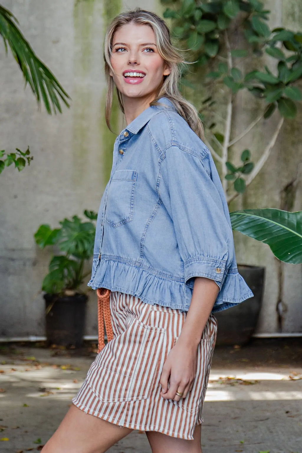 Woman wearing a denim jacket and striped skirt standing outdoors with plants in the background