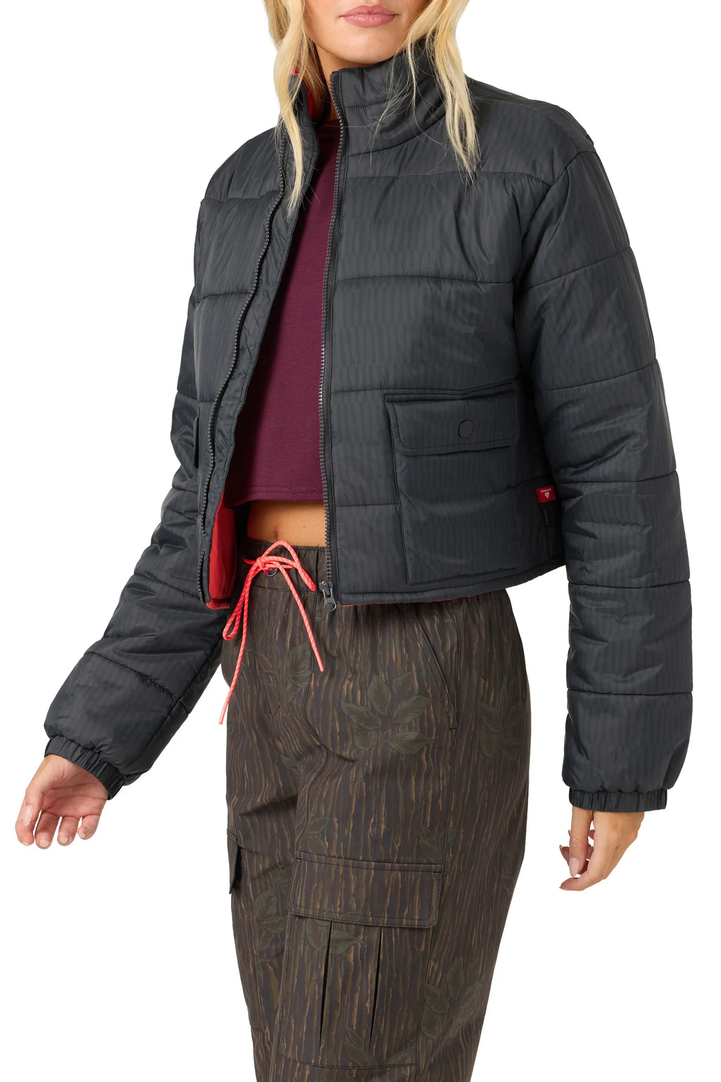 A model wearing a cropped black quilted puffer jacket with a stand-up collar, paired with brown patterned cargo pants and a dark purple crop top, against a solid white background.
