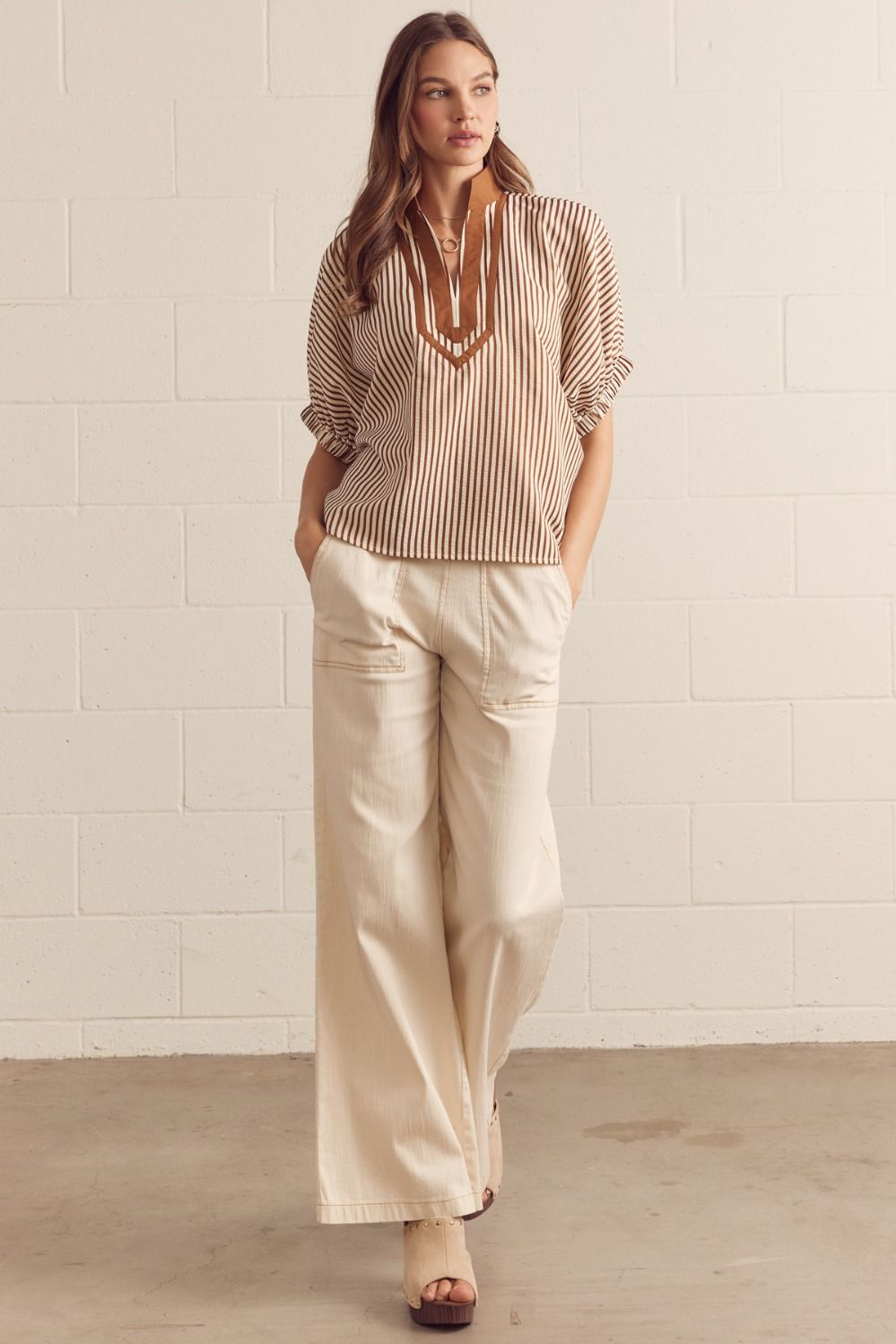 Woman wearing a striped blouse and beige pants against a white tiled wall.