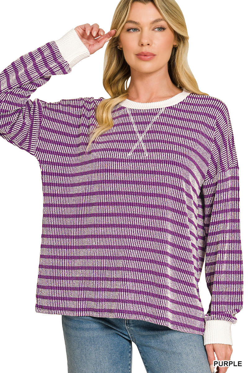 A woman wearing a purple and white horizontally striped knit long-sleeve sweater with white ribbed cuffs and collar, paired with blue jeans against a white background.