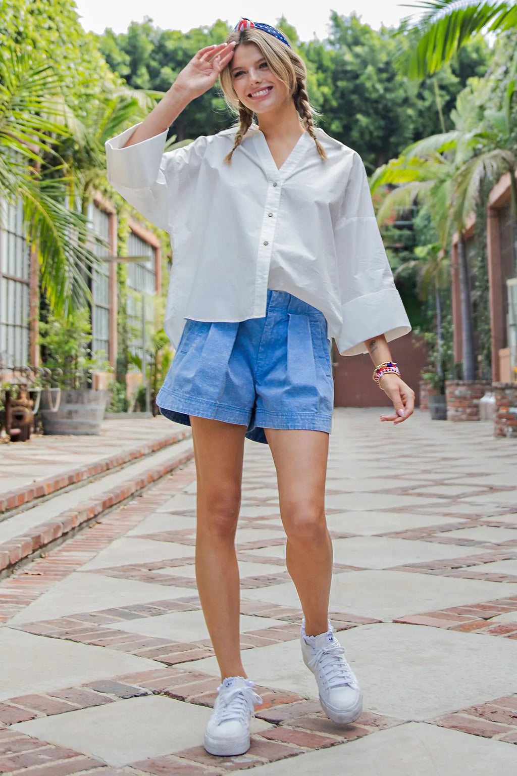 Woman in white shirt and blue shorts standing on a paved walkway with greenery in the background