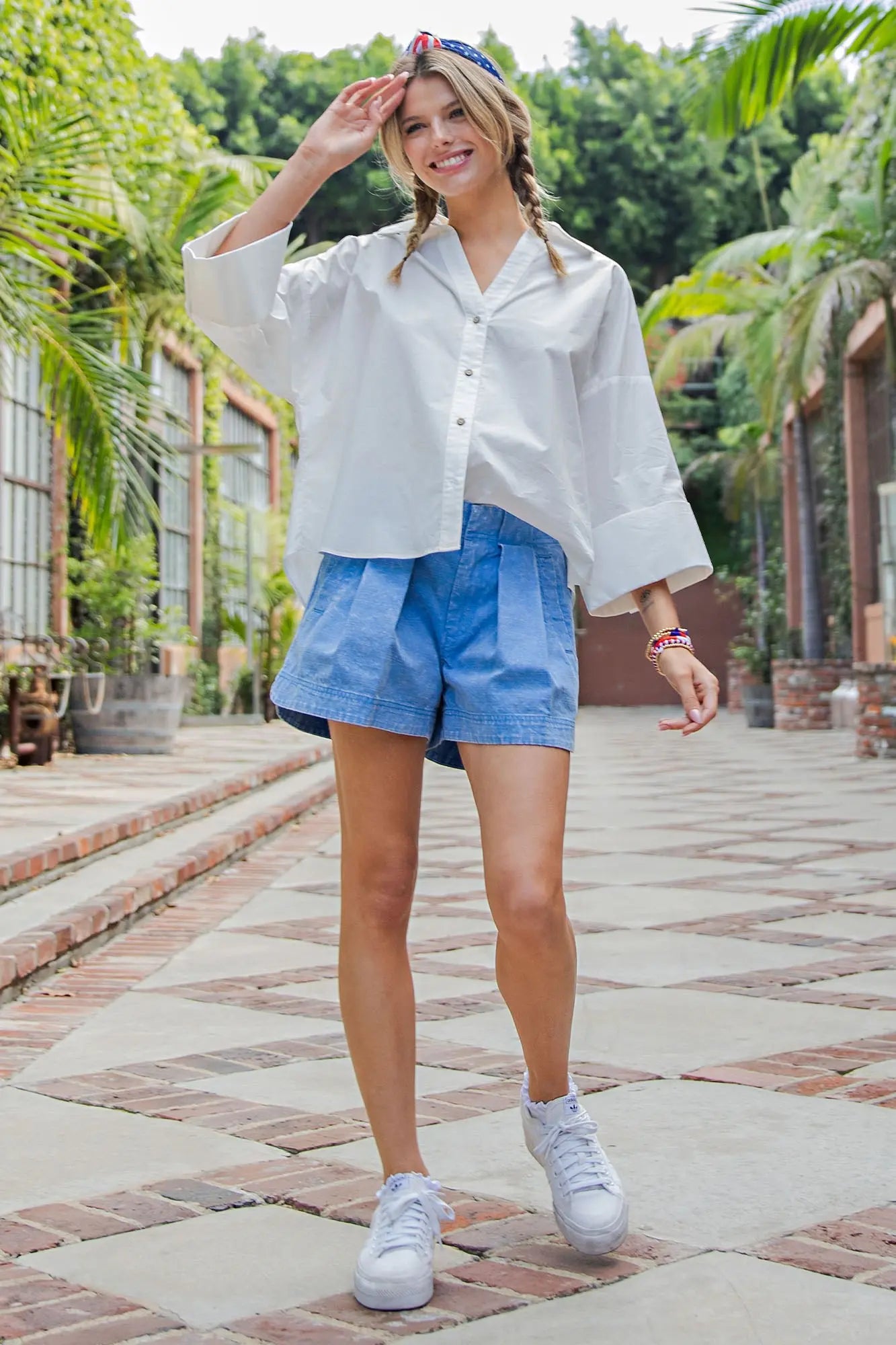Woman in white shirt and blue shorts standing on a paved walkway with greenery in the background