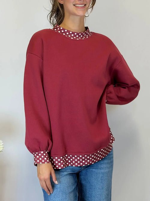 A woman wearing a long-sleeve crewneck sweatshirt in burgundy with contrasting red and white polka dot trim at the collar, cuffs, and hem, paired with blue jeans.