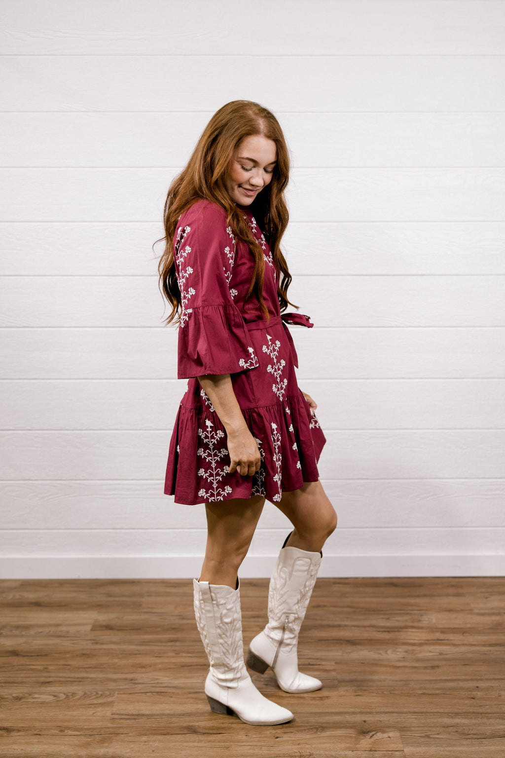 A woman modeling a short burgundy dress with white floral patterns and white cowboy boots, standing against a white shiplap wall on a wooden floor.