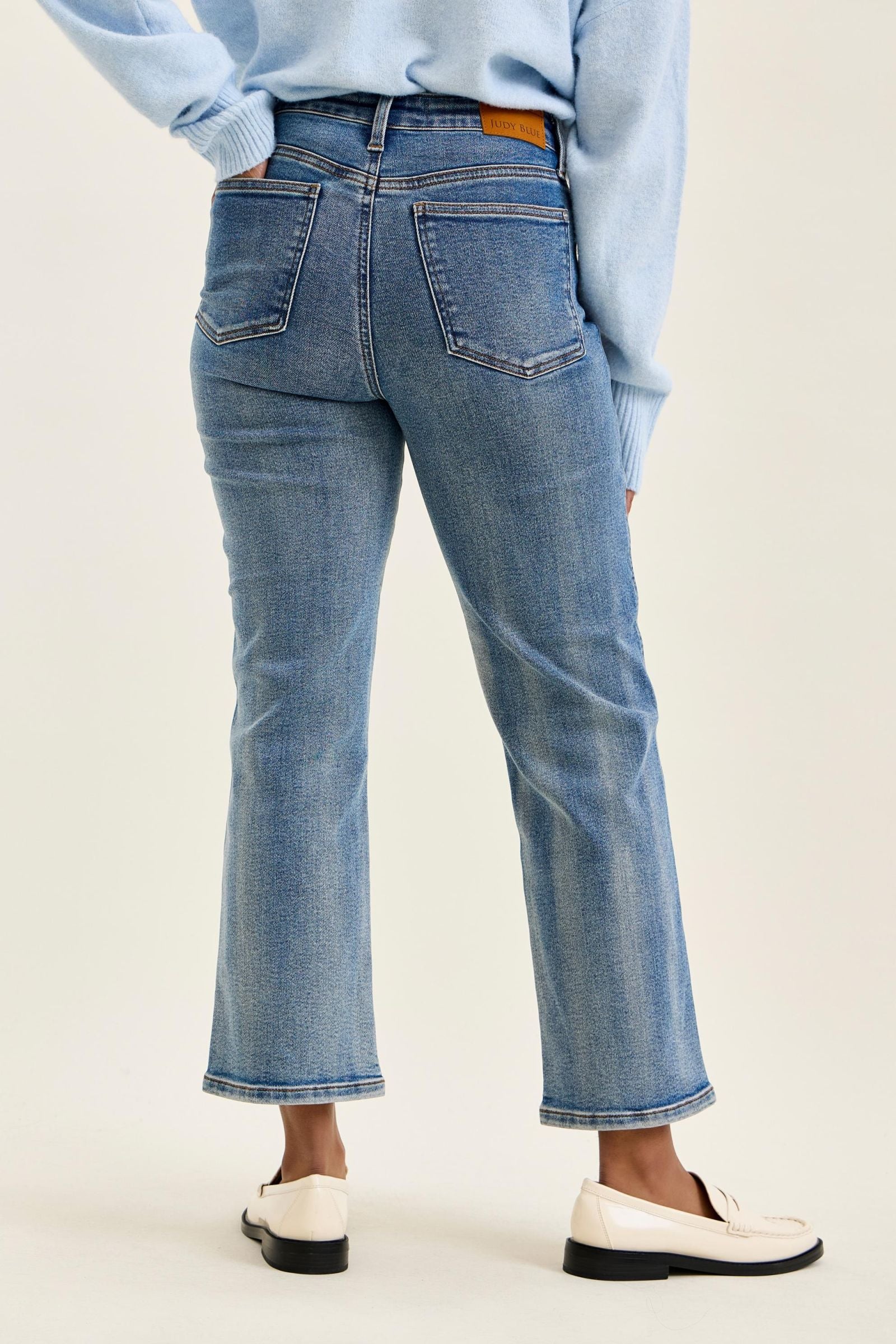 Rear view of a person wearing light blue cropped flare jeans with a light blue sweater and white loafers, against a plain light beige studio background.