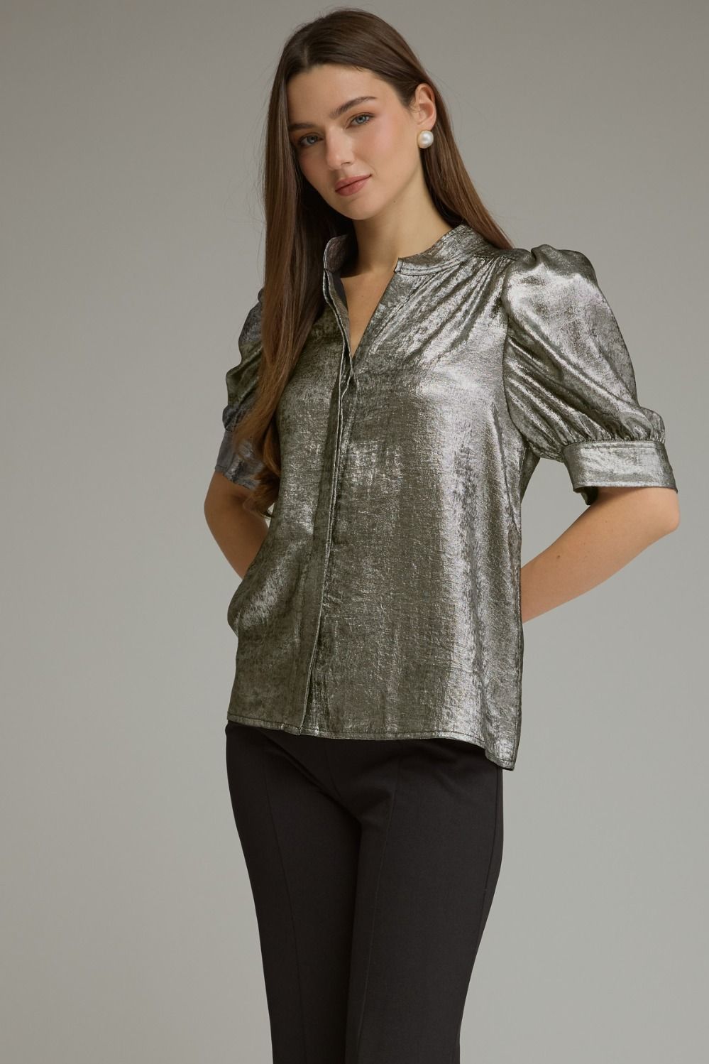 A woman modeling a metallic silver short-sleeve blouse with puffed sleeves and a stand collar, paired with black pants, against a solid gray studio background.