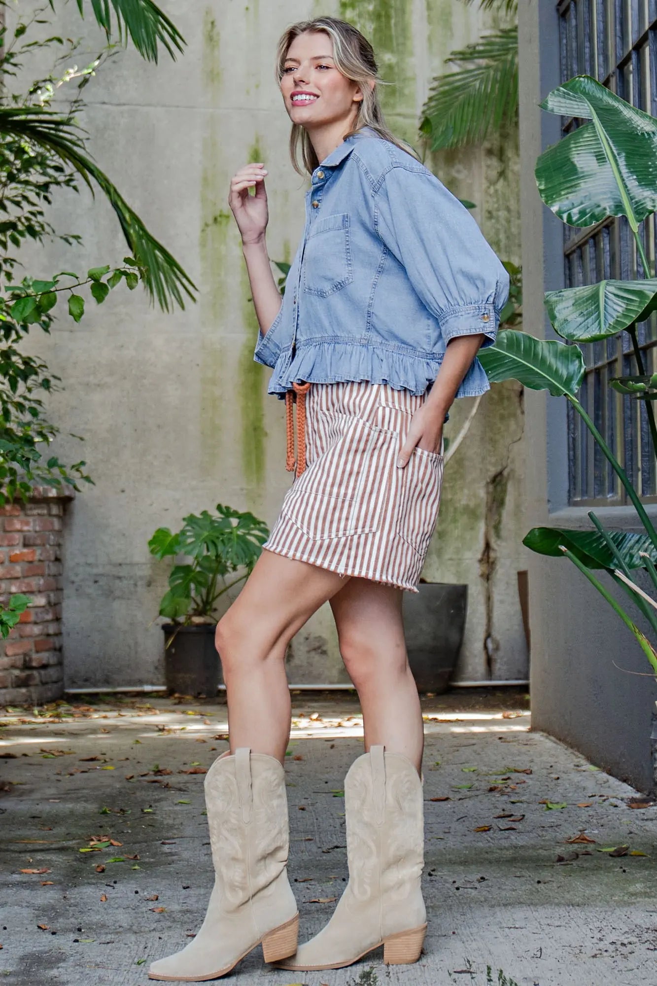 Woman wearing a denim jacket, striped skirt, and beige boots standing outdoors with plants in the background.