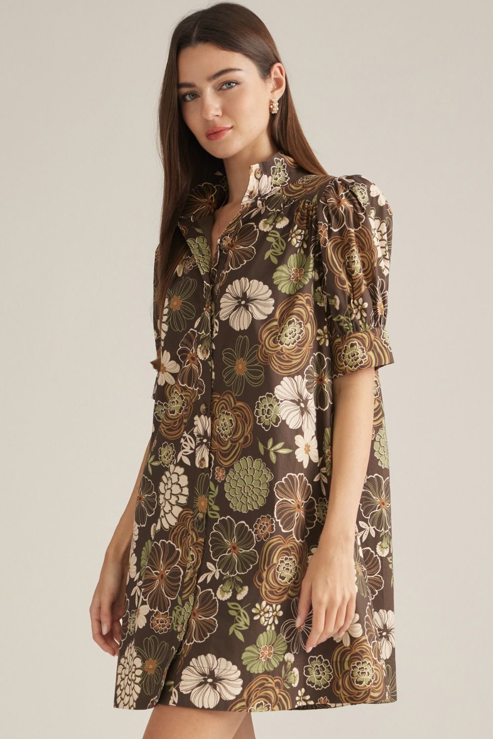 A woman modeling a brown short-sleeve mini dress with a vintage floral and botanical print in beige, green, and brown tones, set against a plain light gray studio background.