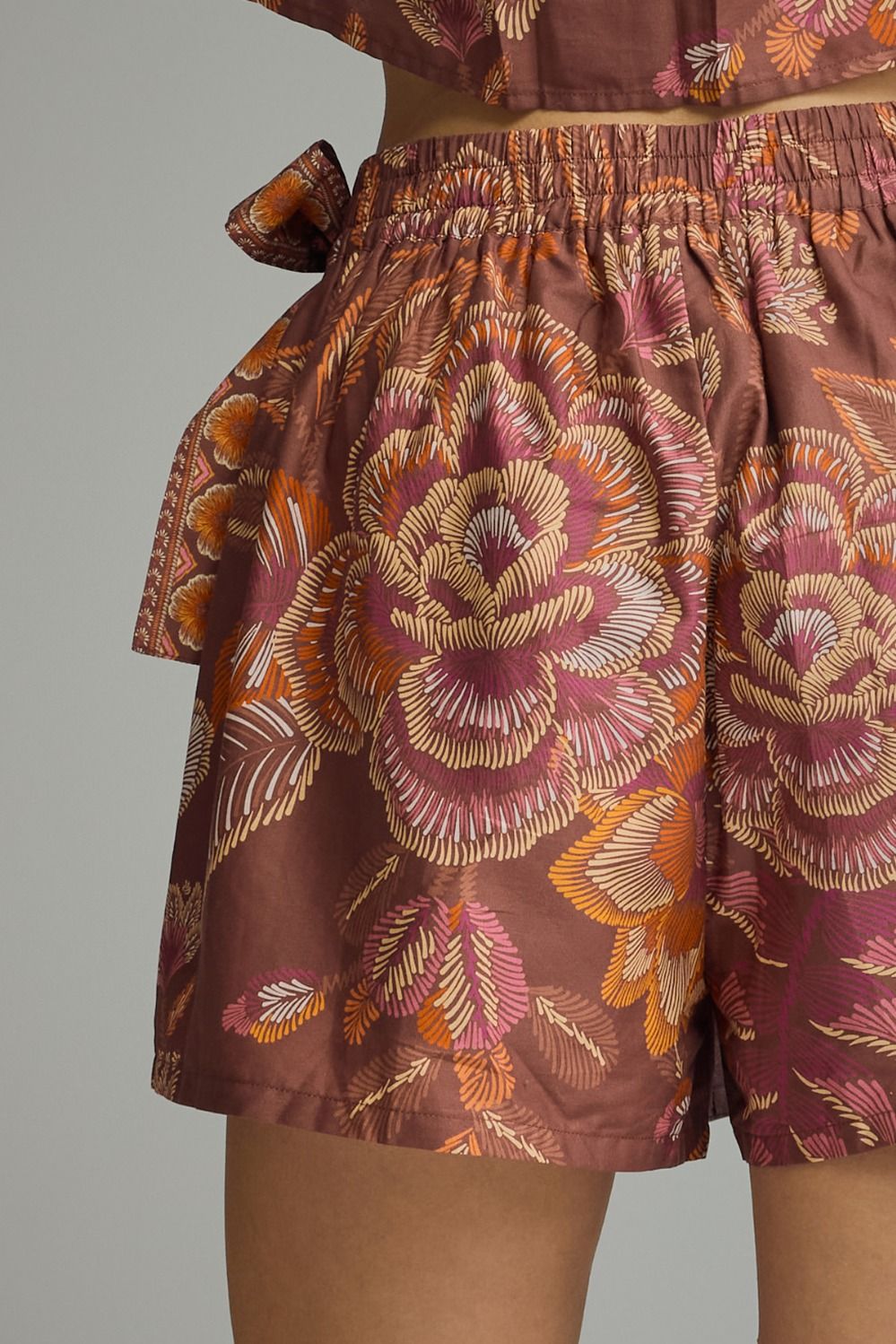 Close-up of a person wearing patterned shorts and a matching top, featuring a large floral print in dusty rose, orange, and cream on a mauve-brown background.
