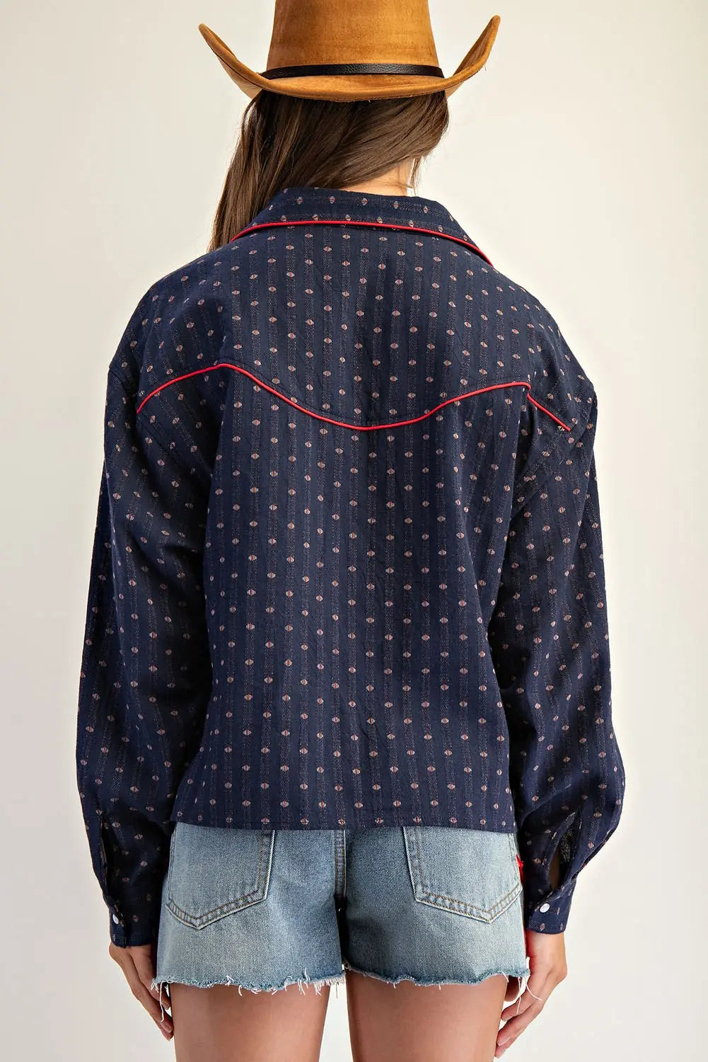Person wearing a navy blue patterned shirt with red accents and denim shorts, standing against a plain background.