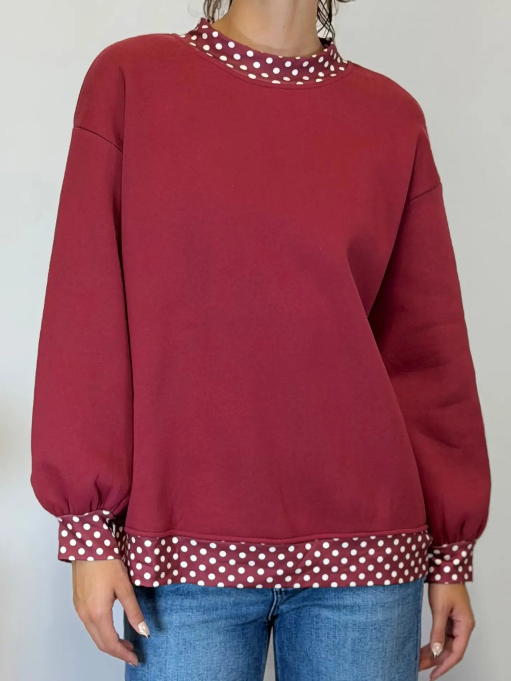 A person wearing a long-sleeve crewneck sweatshirt in deep burgundy with contrasting white polka dot trim at the collar, cuffs, and hem, paired with blue jeans.