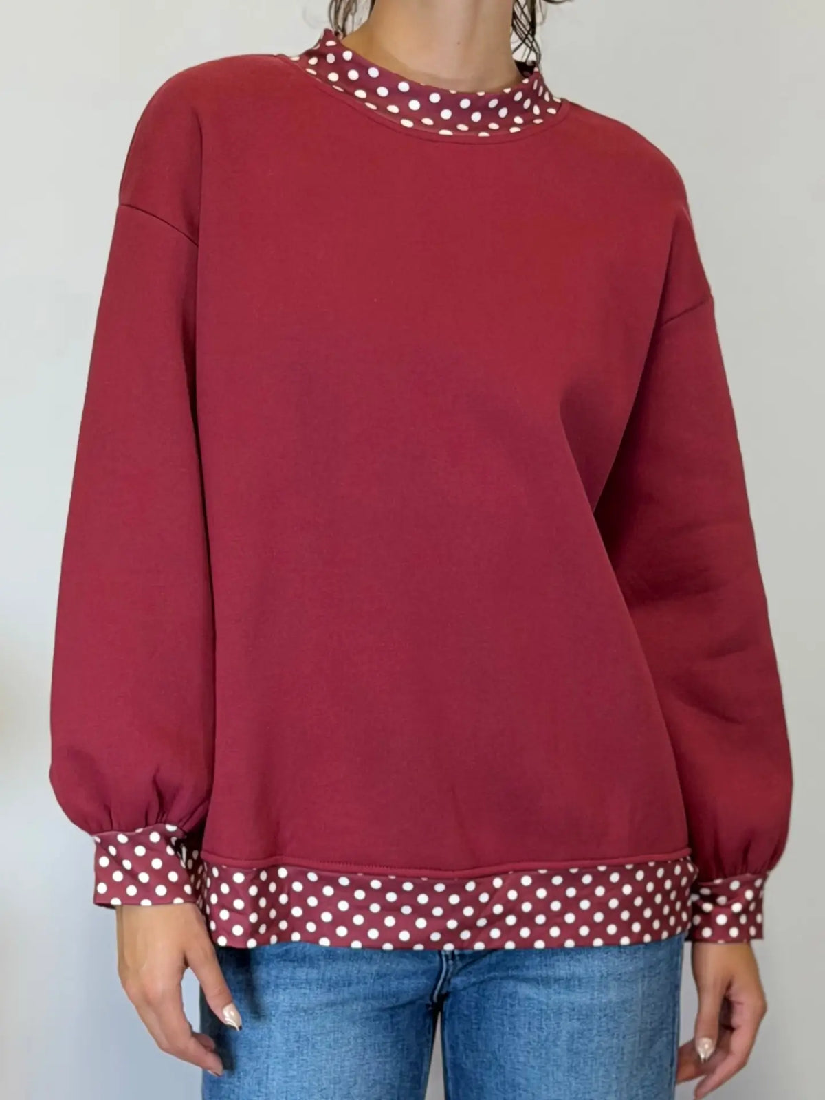 A person wearing a long-sleeve crewneck sweatshirt in deep burgundy with contrasting white polka dot trim at the collar, cuffs, and hem, paired with blue jeans.