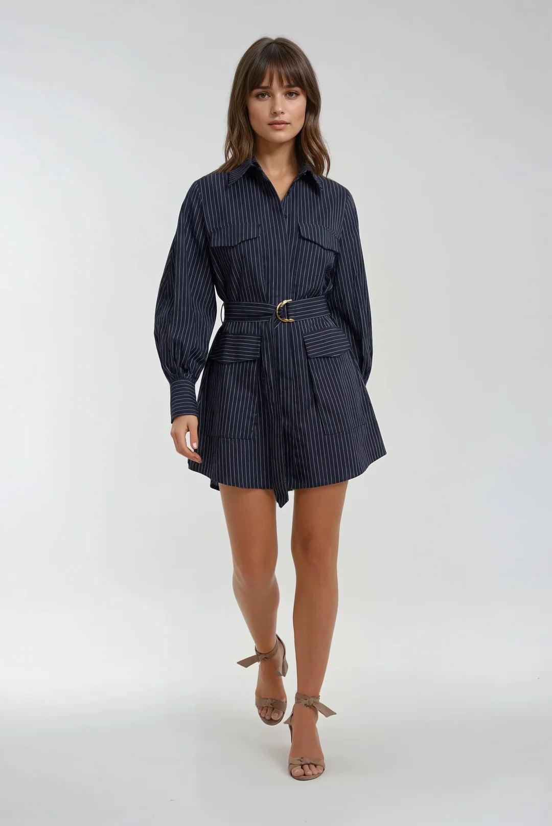 A woman modeling a navy blue and white pinstriped shirt dress with a matching belt and gold buckle, paired with brown strappy high-heeled sandals, against a light gray studio background.