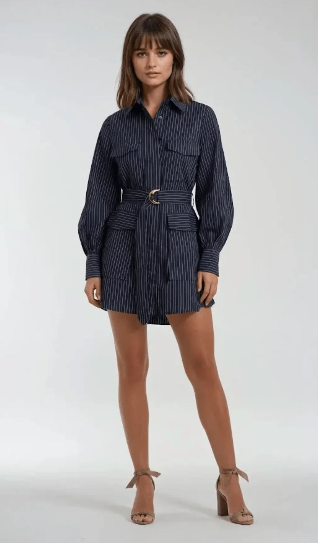 A woman modeling a navy blue and white vertically striped belted shirt dress with long balloon sleeves, a button-up collar, and front flap pockets, paired with beige ankle-strap high heels against a light gray studio background.
