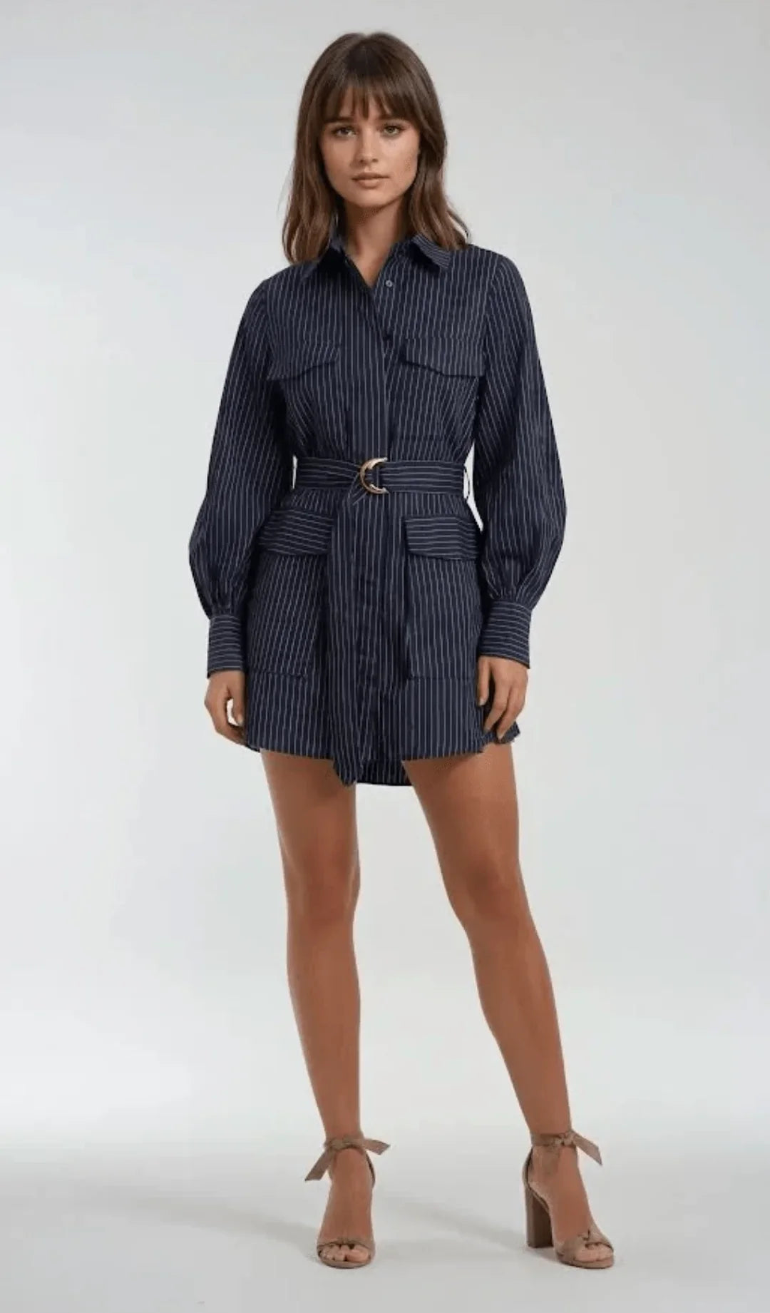 A woman modeling a navy blue and white vertically striped belted shirt dress with long balloon sleeves, a button-up collar, and front flap pockets, paired with beige ankle-strap high heels against a light gray studio background.