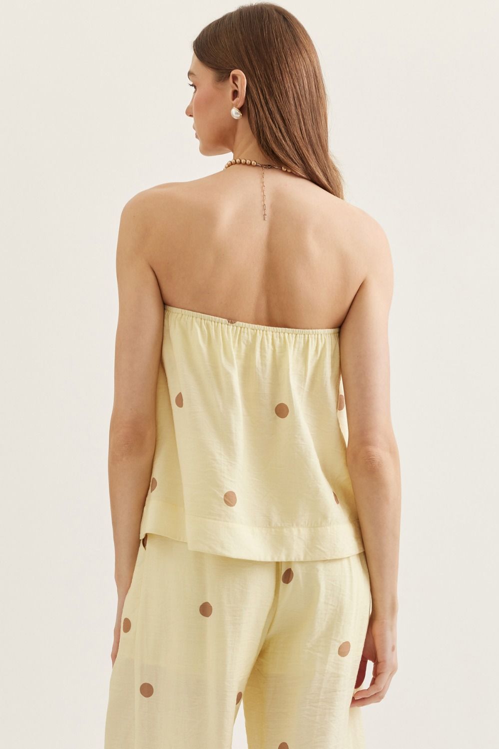 Woman wearing a light yellow strapless top and pants with brown buttons on a beige background