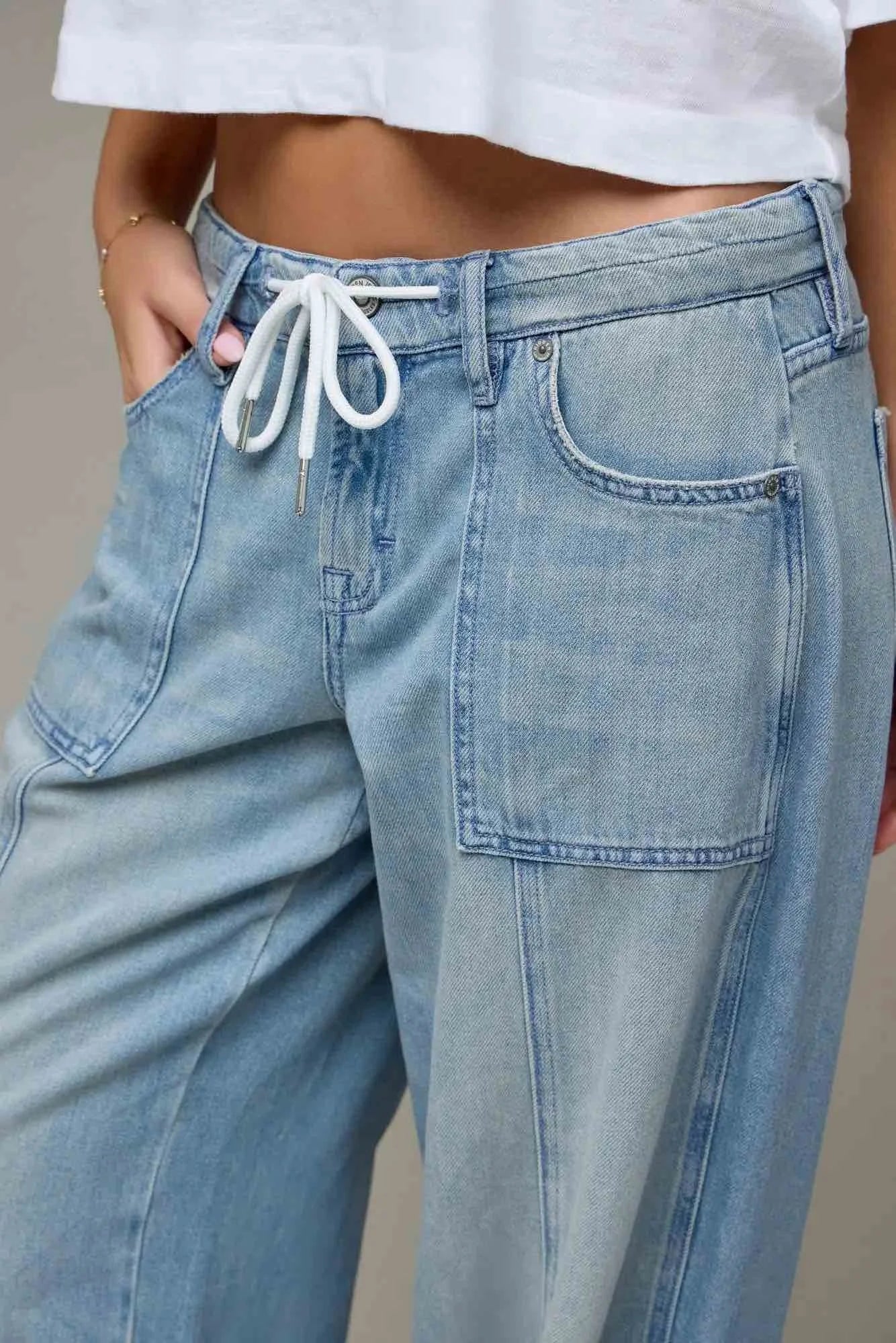 Close-up of a person wearing light blue wide-leg denim jeans with a white drawstring waistband, paired with a white cropped top against a neutral beige background.