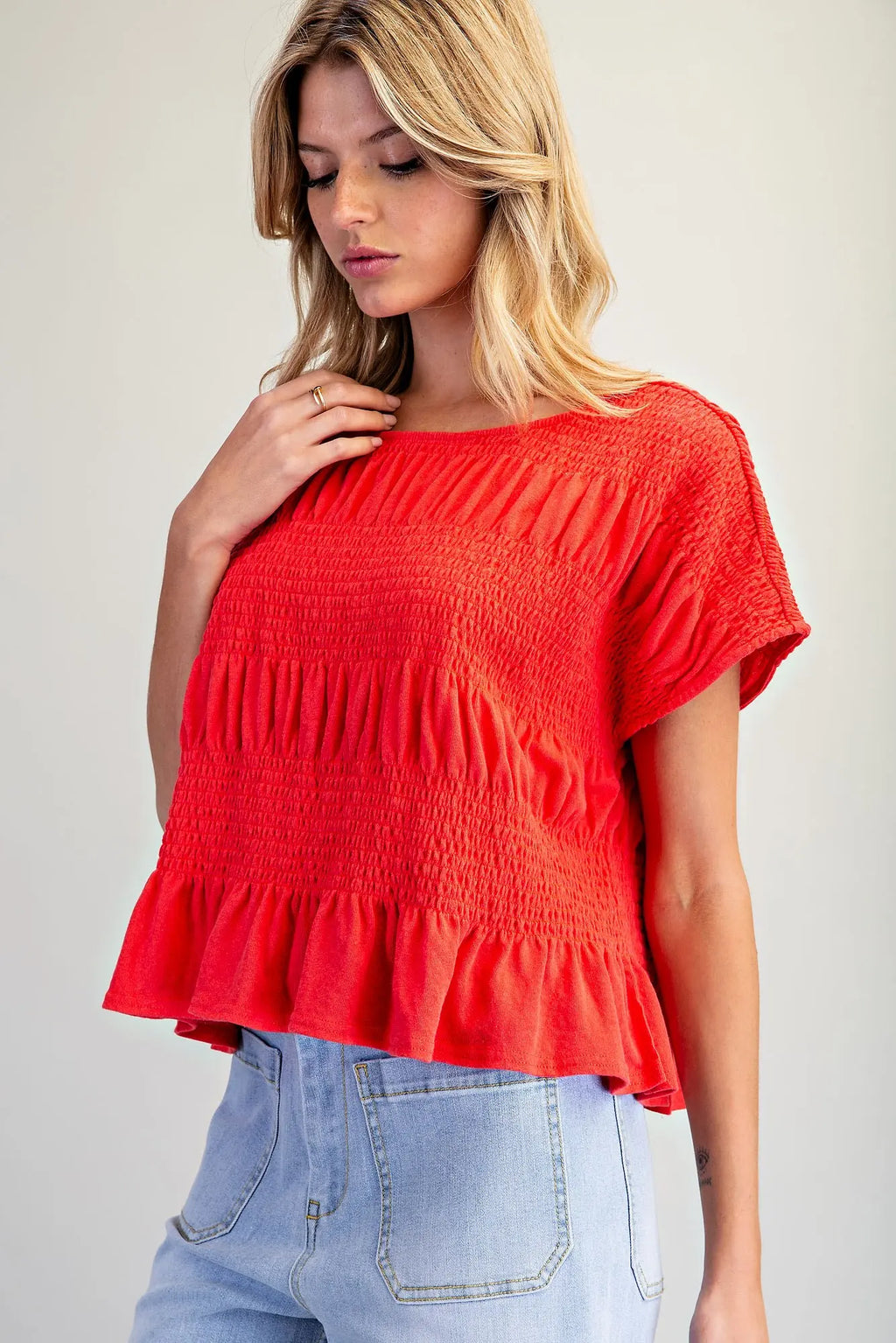 A woman wearing a bright red short-sleeve top with a smocked bodice and ruffled hem, paired with light blue jeans, photographed against a plain light gray background.