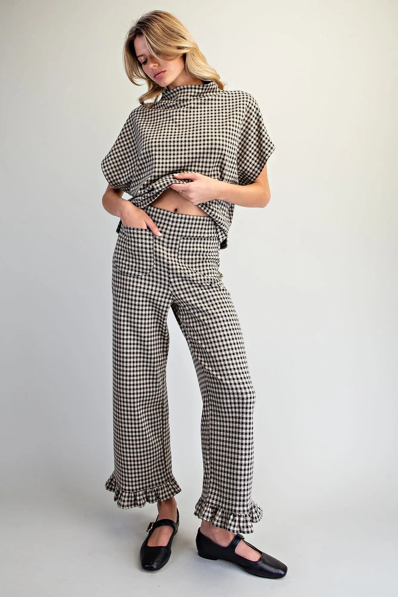 A woman modeling a matching black and white gingham two-piece set consisting of a short-sleeve mock-neck top and wide-leg ruffled pants, paired with black Mary Jane shoes, against a plain light gray studio background.