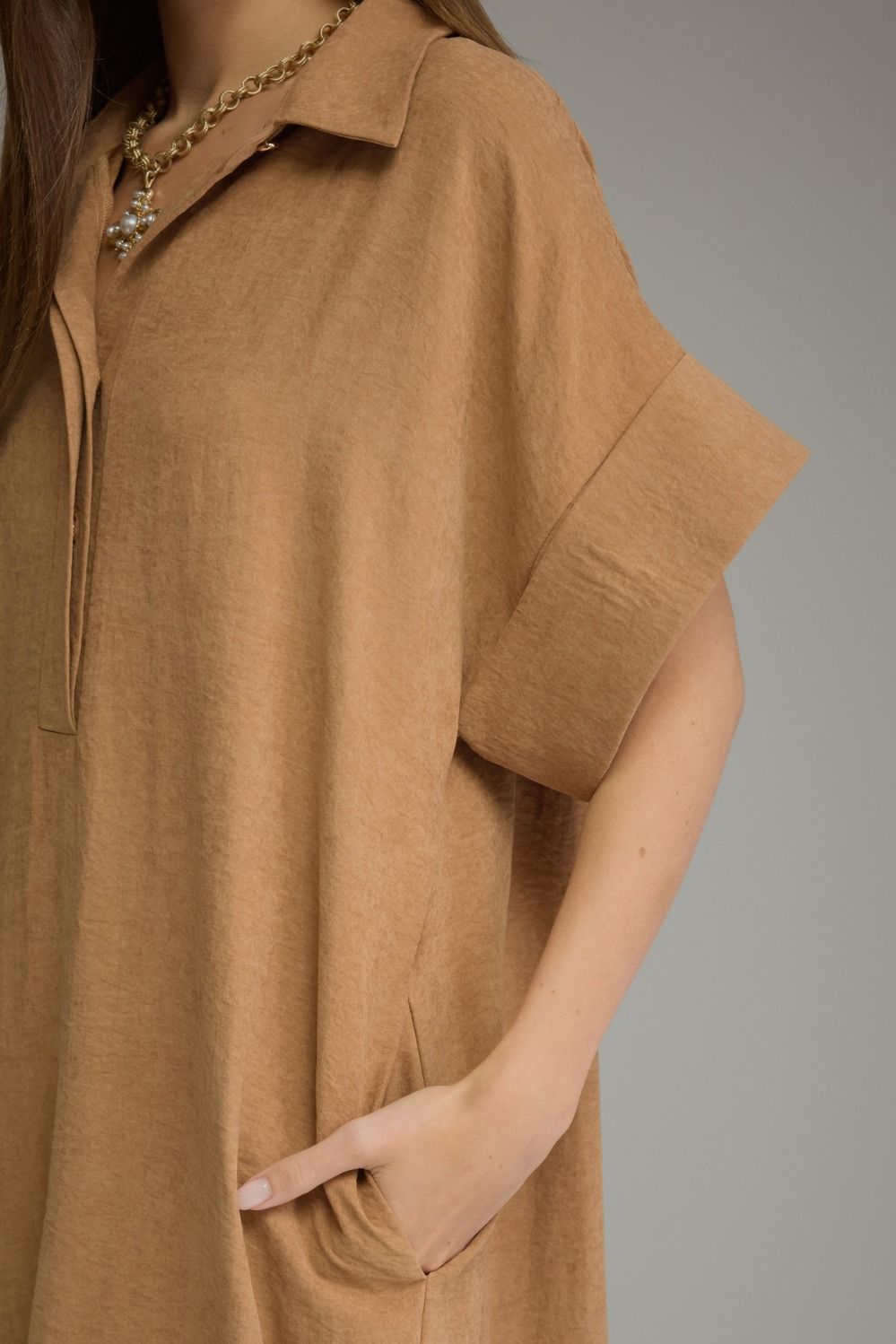 A close-up of a woman wearing a loose, short-sleeve collared tunic dress in a warm camel brown color, paired with a gold chain necklace, against a solid gray background.