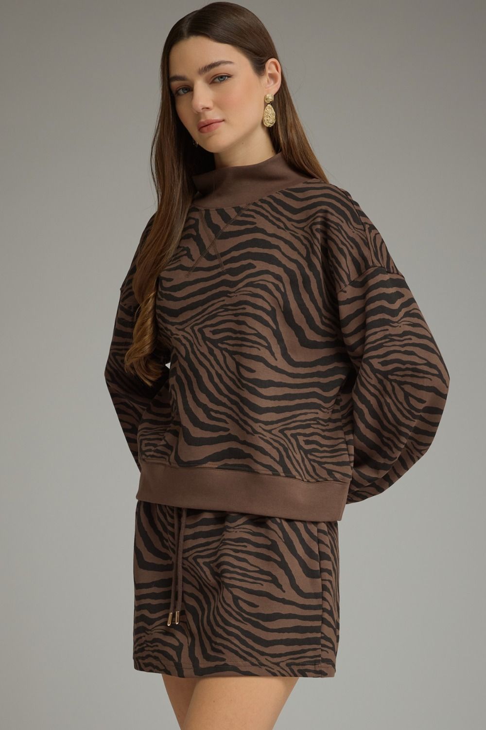 A woman modeling a brown and black zebra-print knit set consisting of a mock-neck long-sleeve sweatshirt and a matching mini skirt, against a solid gray studio background.