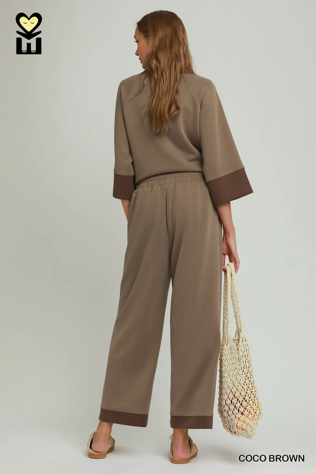 Person wearing a brown outfit holding a woven bag on a plain background