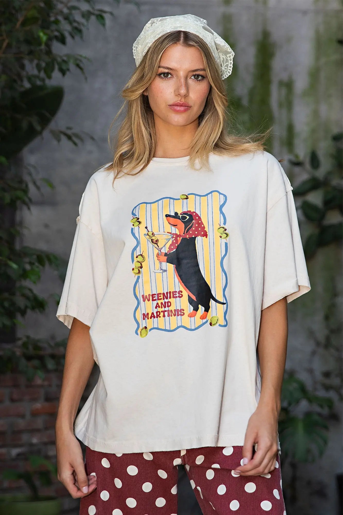 A woman wearing an oversized cream-colored graphic t-shirt with a 'Weenie and Martini' dachshund print, styled with polka-dot pants and a lace headband in an outdoor setting.