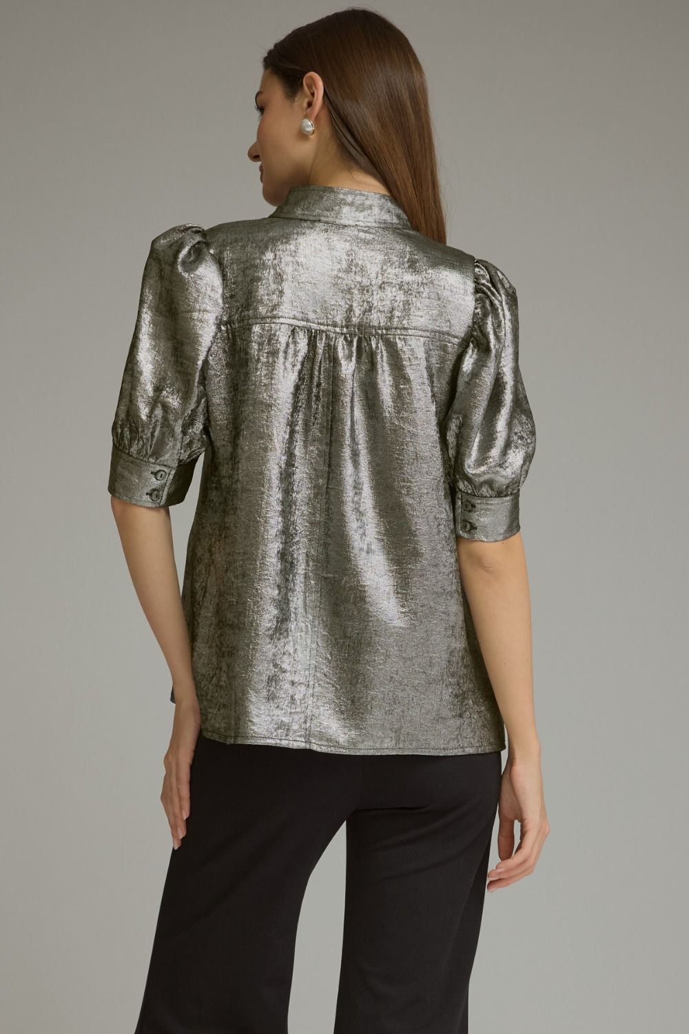 A woman shown from the back wearing a metallic silver short-sleeve blouse with puffed sleeves and black pants, photographed against a plain light gray studio background.