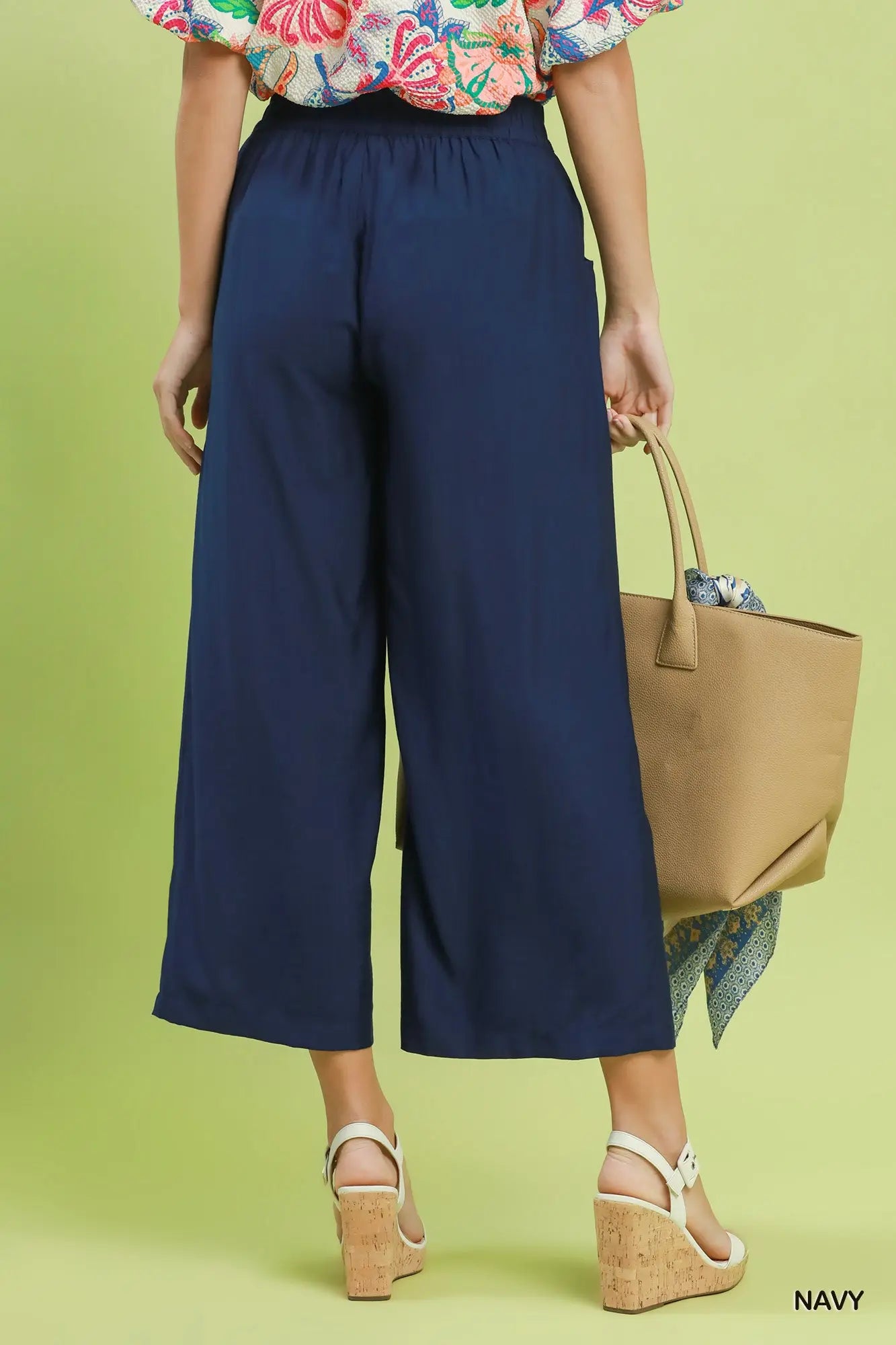 Person wearing navy wide-leg pants with a floral top and beige sandals, holding a tan handbag on a green background.