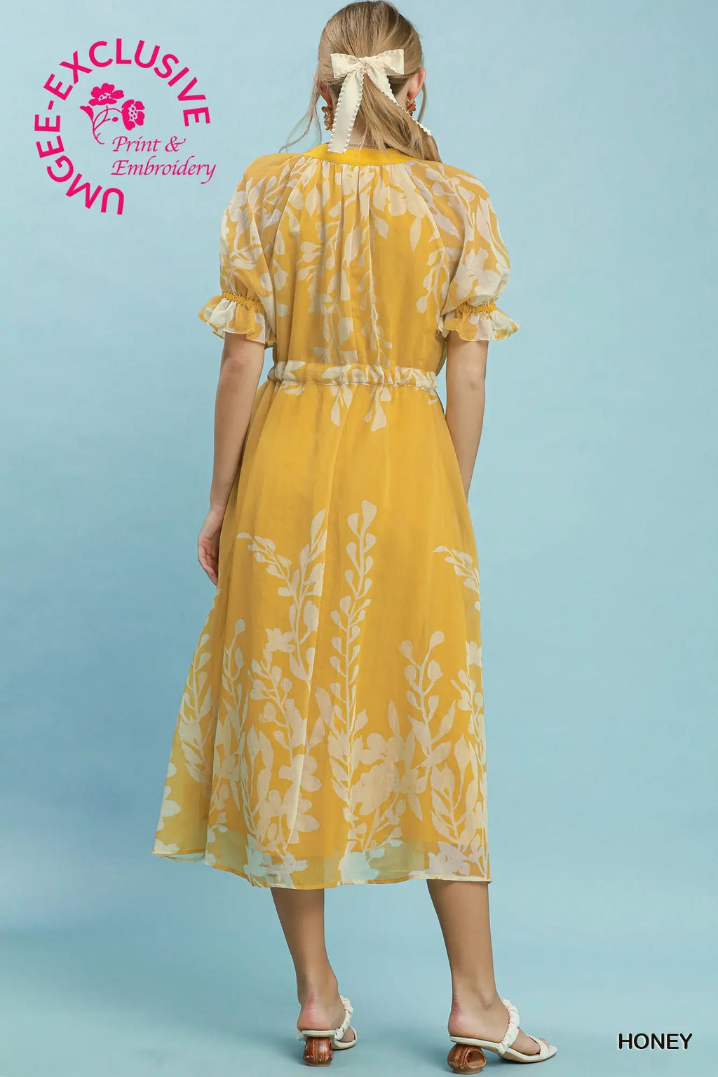 Woman wearing a yellow dress with floral pattern on a light blue background, featuring Umgee Exclusive branding.