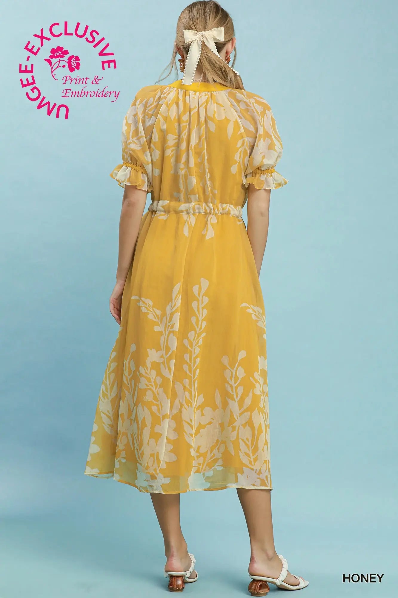 Woman wearing a yellow dress with floral pattern on a light blue background, featuring Umgee Exclusive branding.