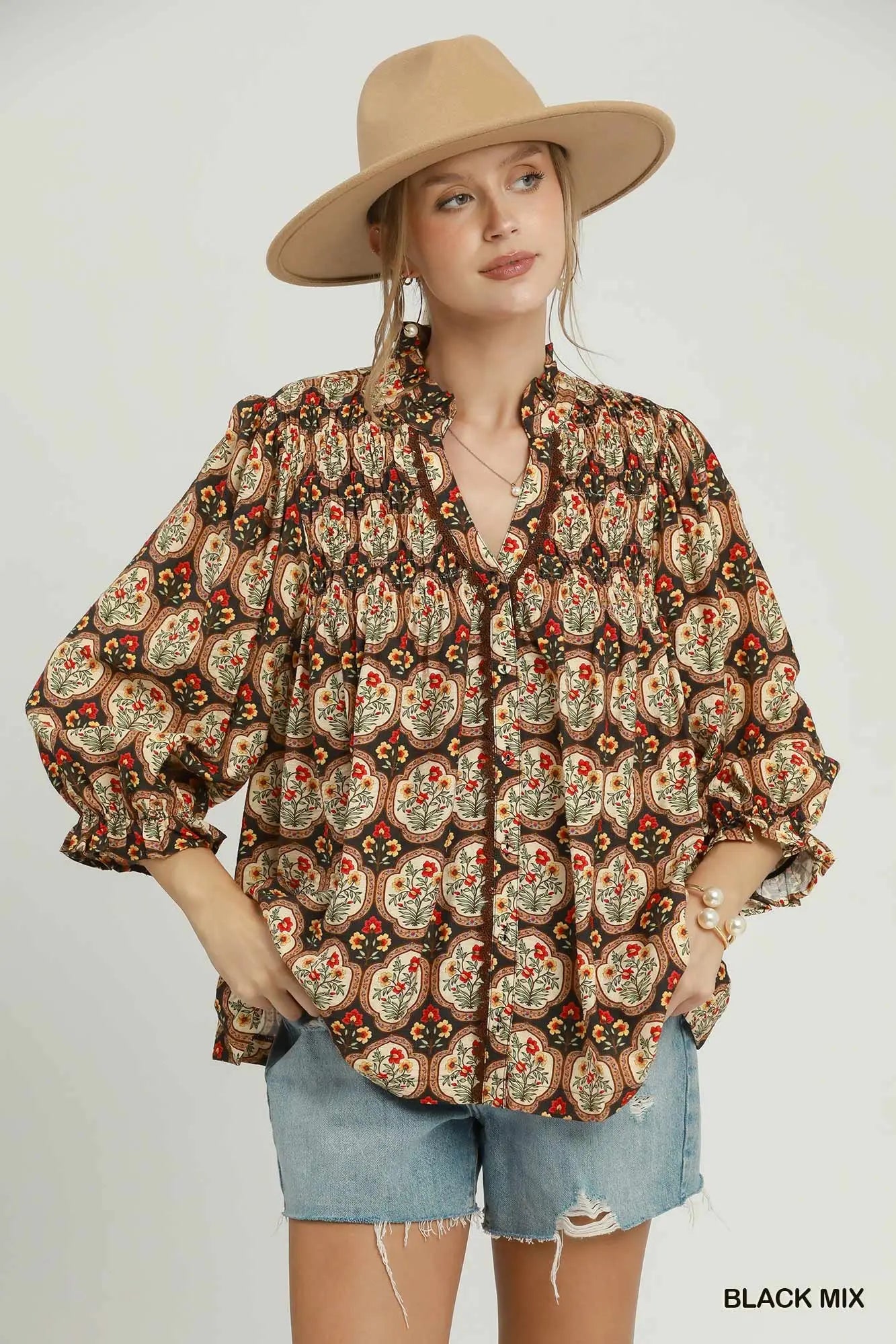 A woman modeling a bohemian-style long-sleeve blouse with a brown, beige, and red paisley pattern, paired with denim shorts and a tan wide-brim hat against a light gray background.