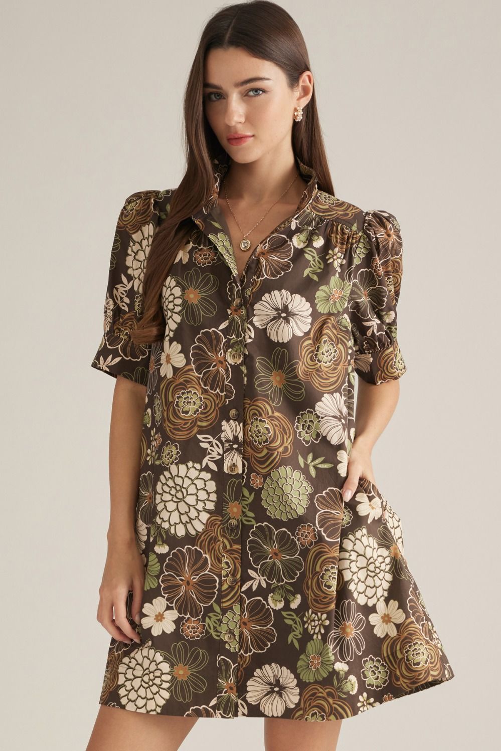 A woman modeling a short-sleeve button-front dress with a brown base and a large floral print in beige, cream, olive green, and brown tones, photographed against a plain light beige studio background.