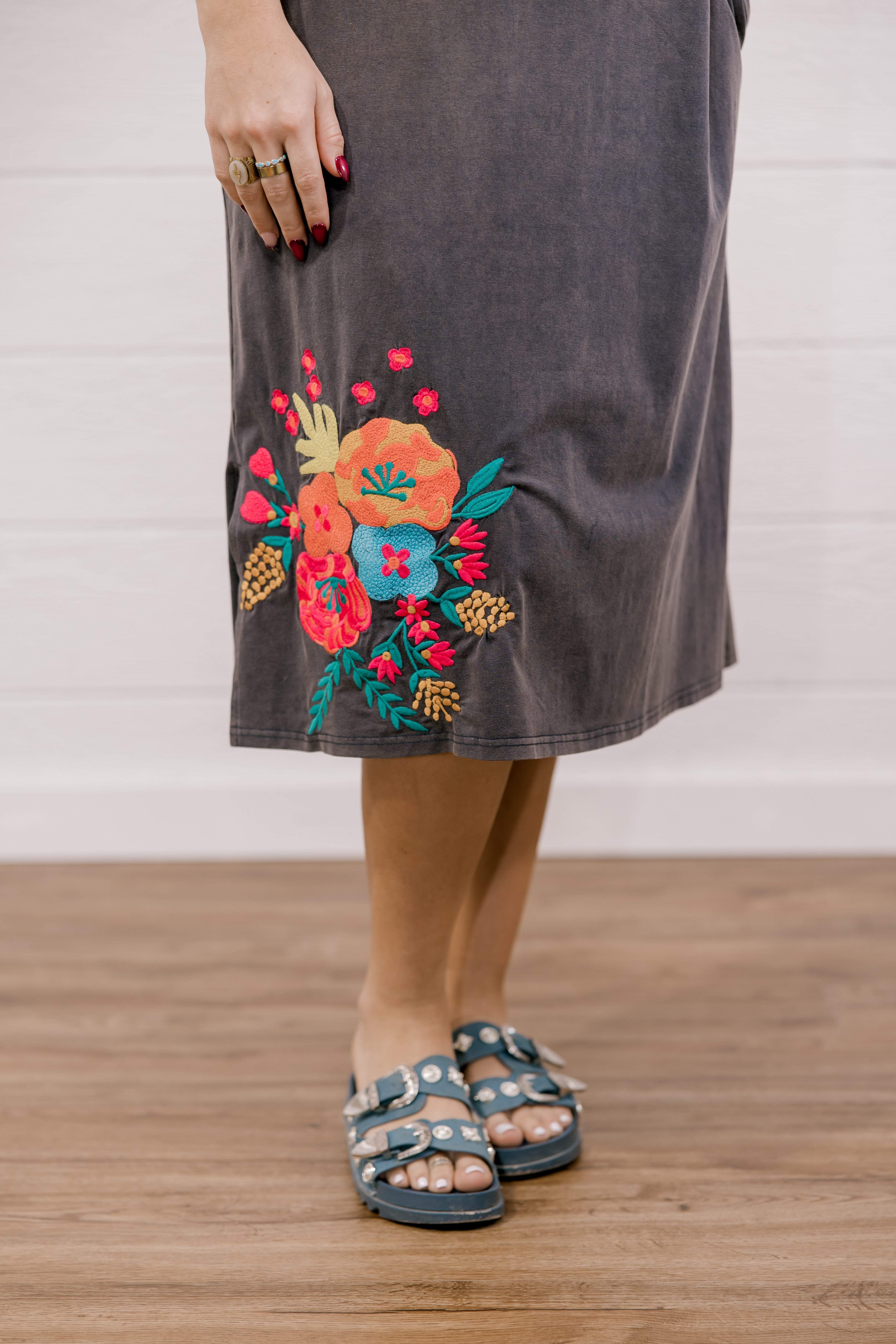 Person wearing a dark gray skirt with colorful floral embroidery on a wooden floor.