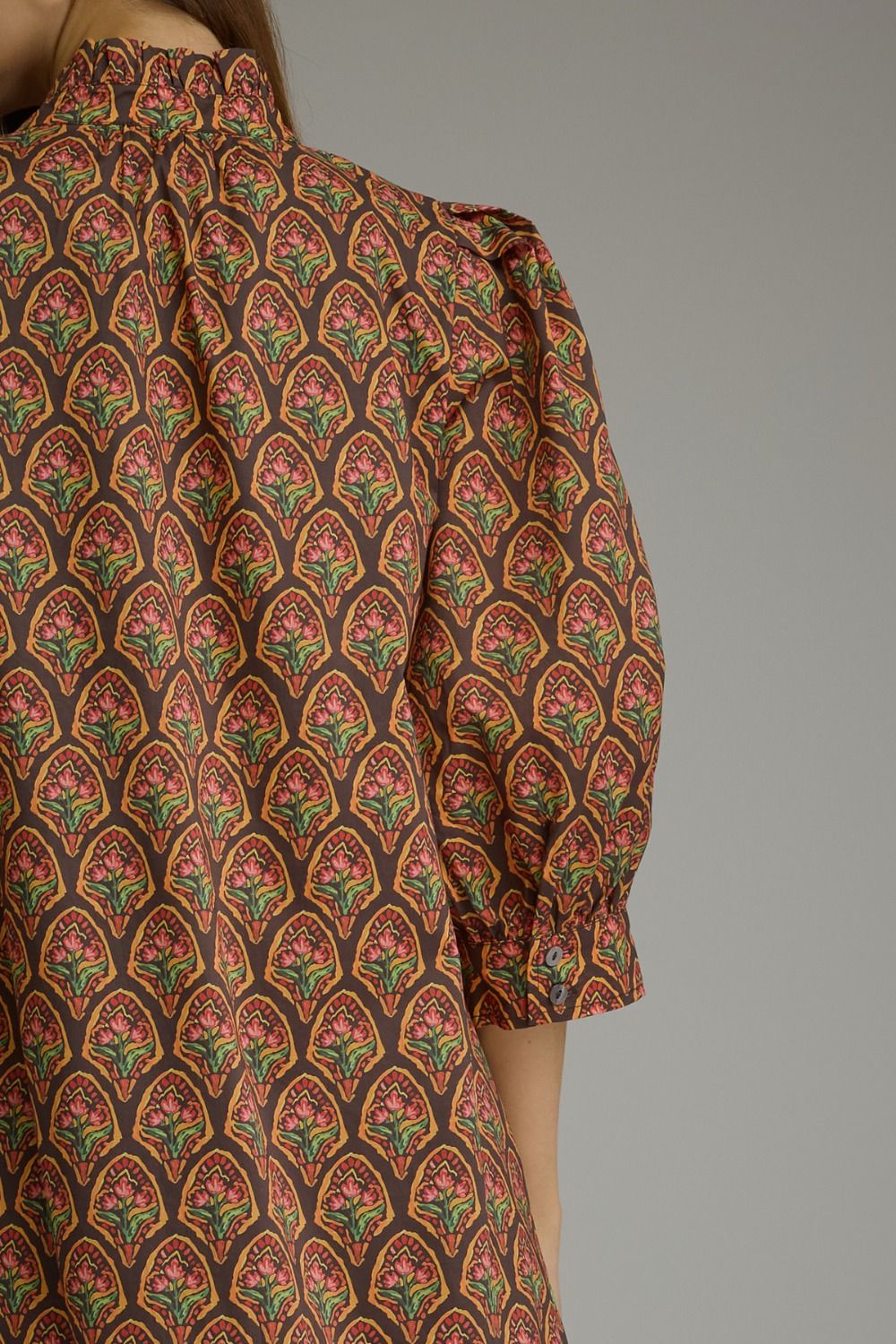 Close-up of a patterned blouse with puff sleeves, featuring a repeating arch motif in rust red, olive green, and gold on a dark brown base, shown on a model against a plain gray background.