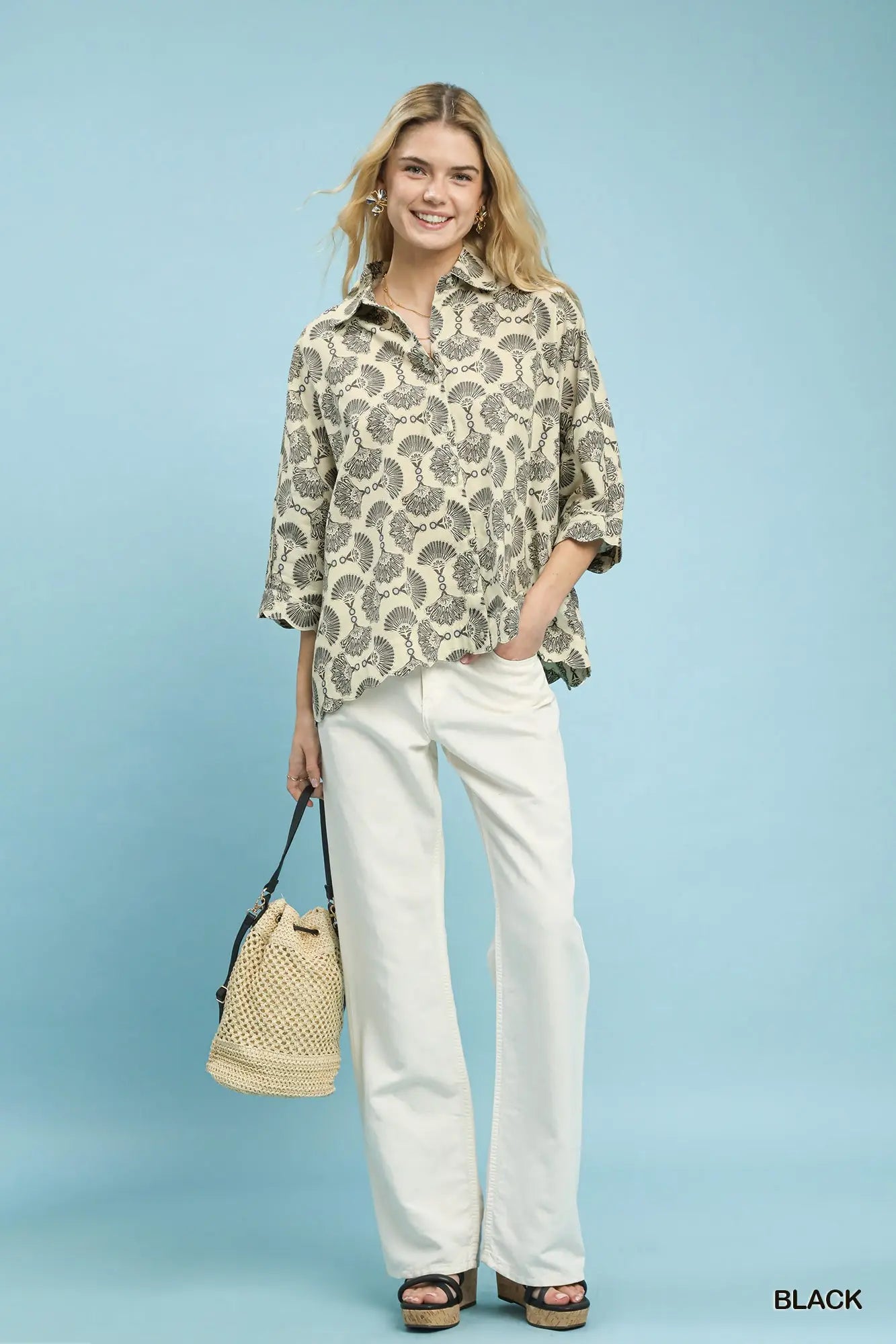 Woman wearing a patterned shirt and white pants with a beige bag against a light blue background