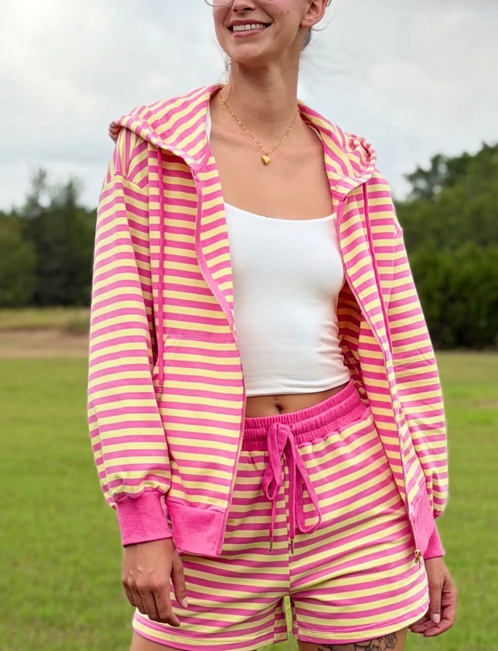 A woman wearing a pink and yellow striped zip-up hoodie and matching striped shorts with a white crop top, standing outdoors in a grassy field.