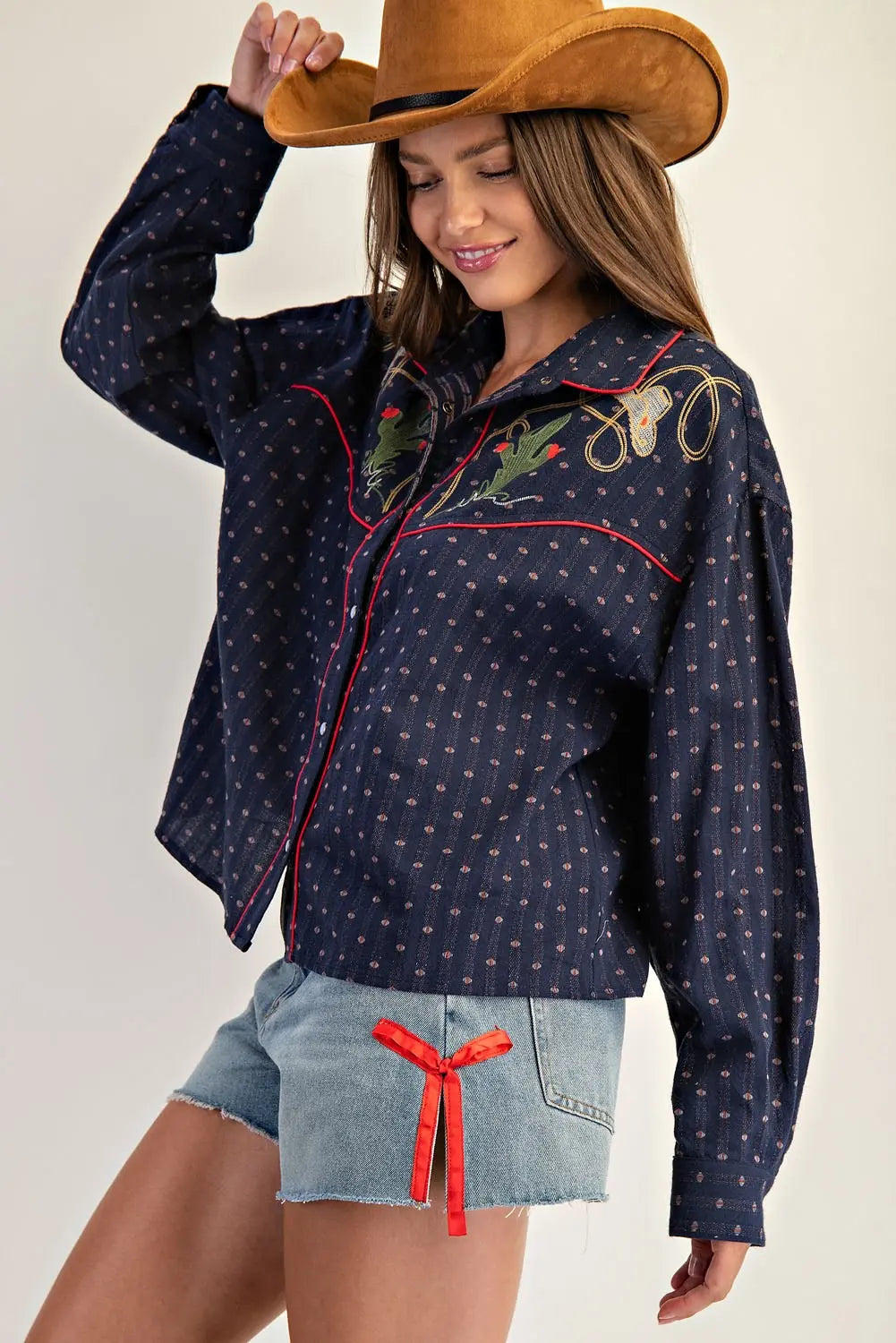 Woman wearing a navy blue embroidered shirt and denim shorts with a cowboy hat.