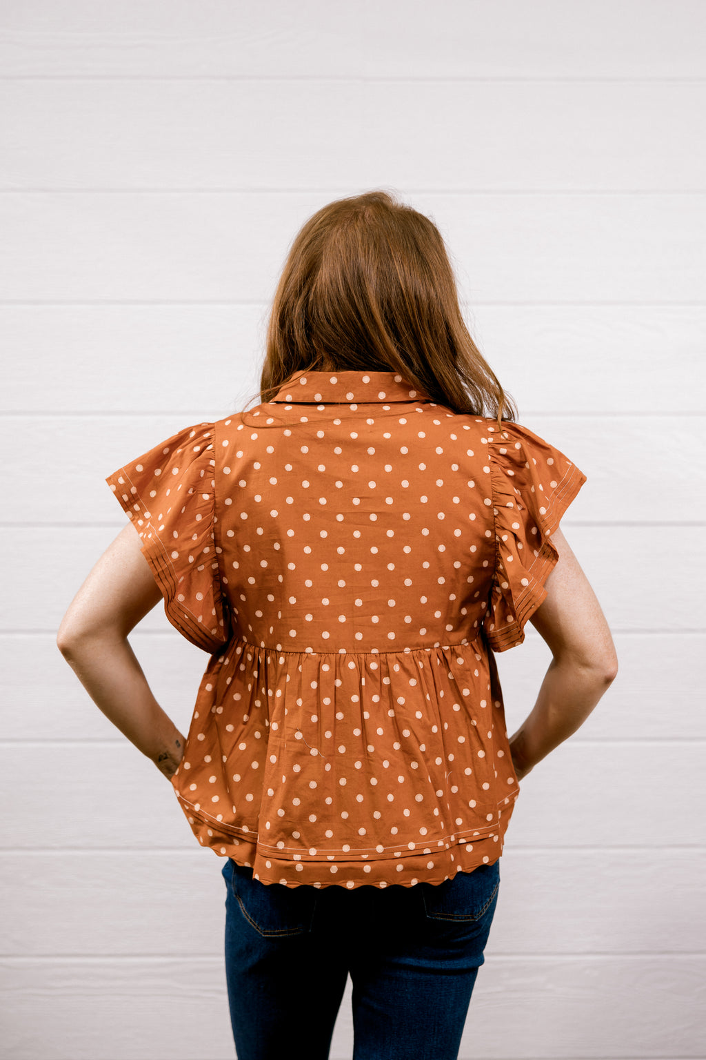 A woman viewed from behind wearing a rust-orange short-sleeve blouse with white polka dots and a gathered peplum waist, paired with dark blue jeans, standing against a white paneled wall.