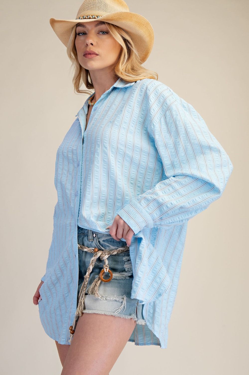 Woman wearing a light blue striped shirt and denim shorts with a beige hat on a beige background